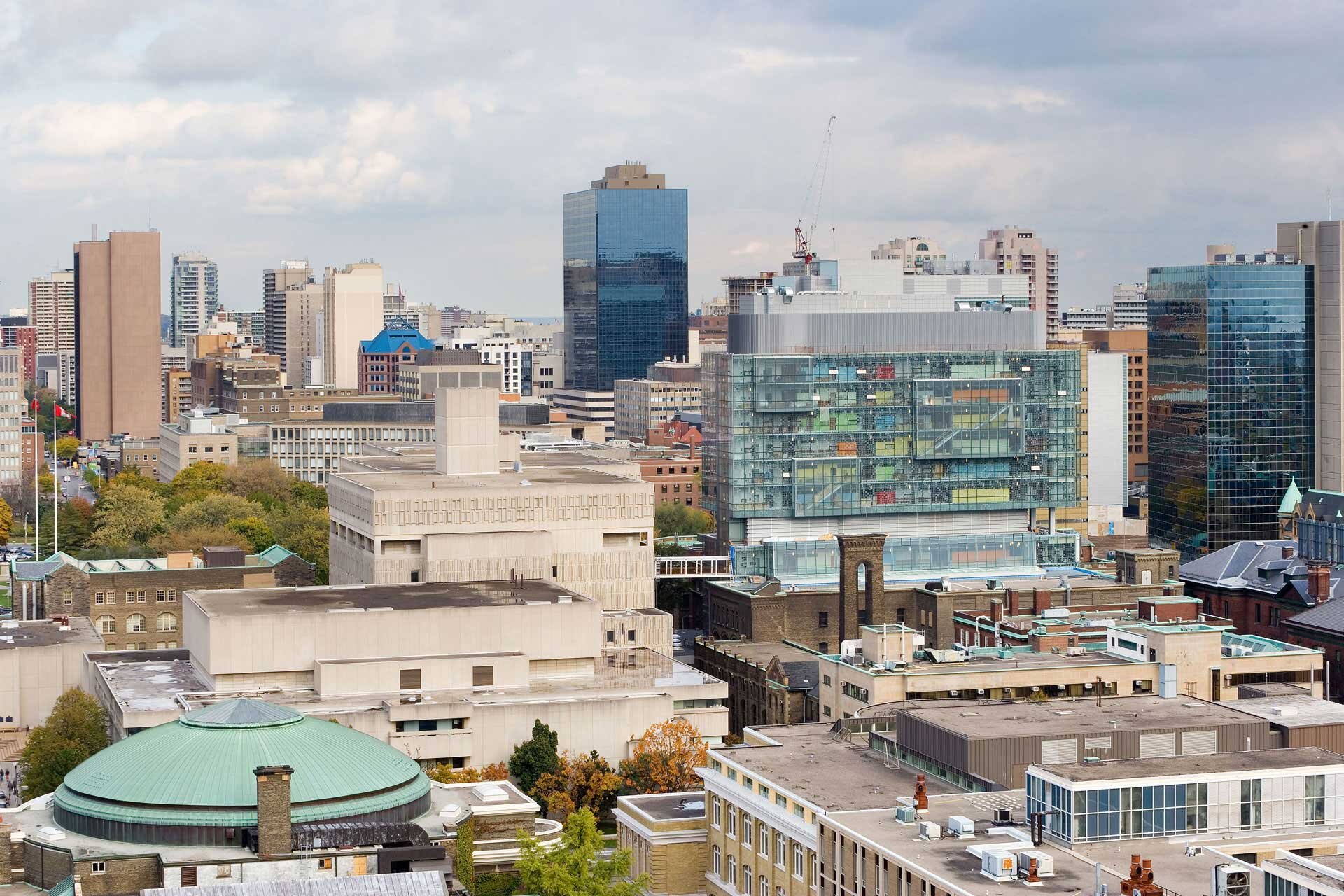 Project by Behnisch Architekturbuero, The Donnelly Centre for Cellular and Biomolecular Research. City skyline with medium-sized and tall buildings, including the Donnelly Center with its glass facade, viewed from above under a cloudy sky.