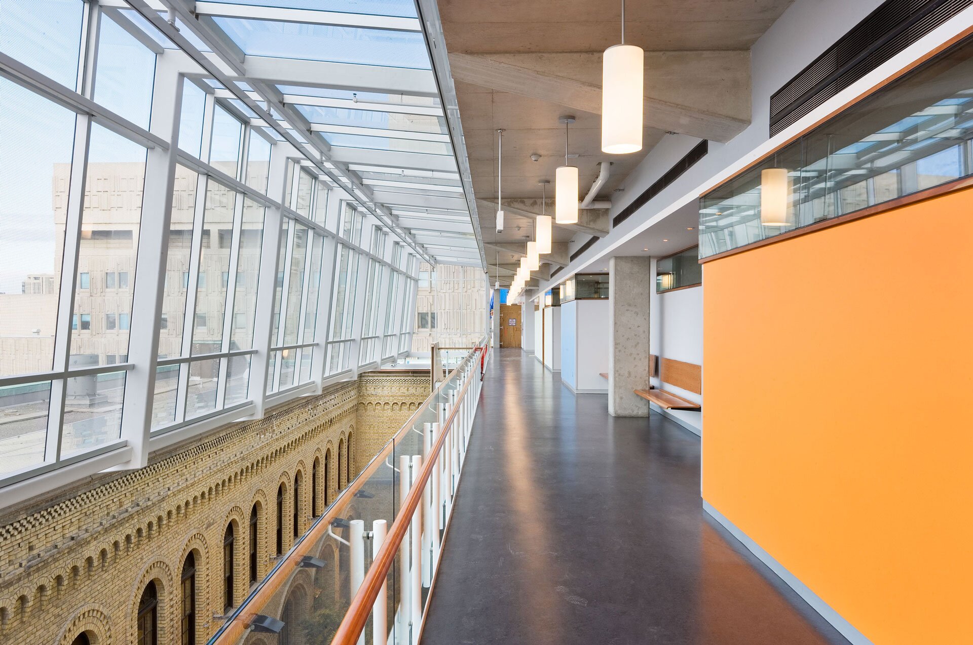 Project by Behnisch Architekturbuero, The Donnelly Centre for Cellular and Biomolecular Research. Interior corridor with glass facade and skylight, overlooking a brick atrium below, with railings, pendant lights and an orange accent wall on the right side of the picture.