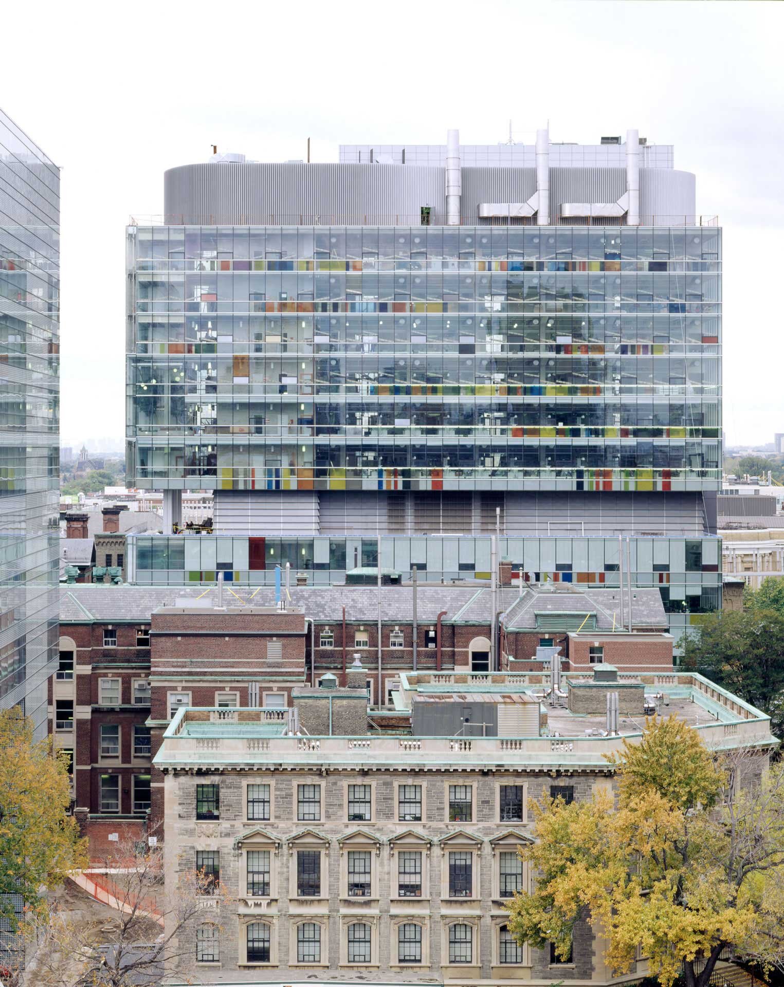 Project by Behnisch Architekturbuero, The Donnelly Centre for Cellular and Biomolecular Research. Front view of the glass office building with visible interior and roof structure, rising above older brick and stone buildings in an urban environment.