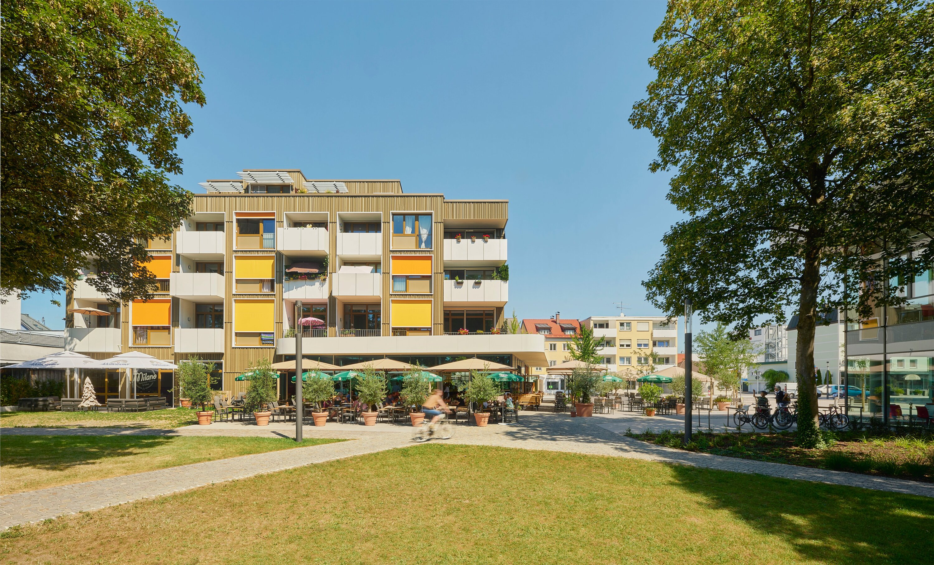 Project by Behnisch Architekturbuero, Mixed-use Building Rathausplatz. Apartment building with balconies, orange and yellow shades, caf&eacute; terrace below, trees and lawn in foreground, and people cycling past.