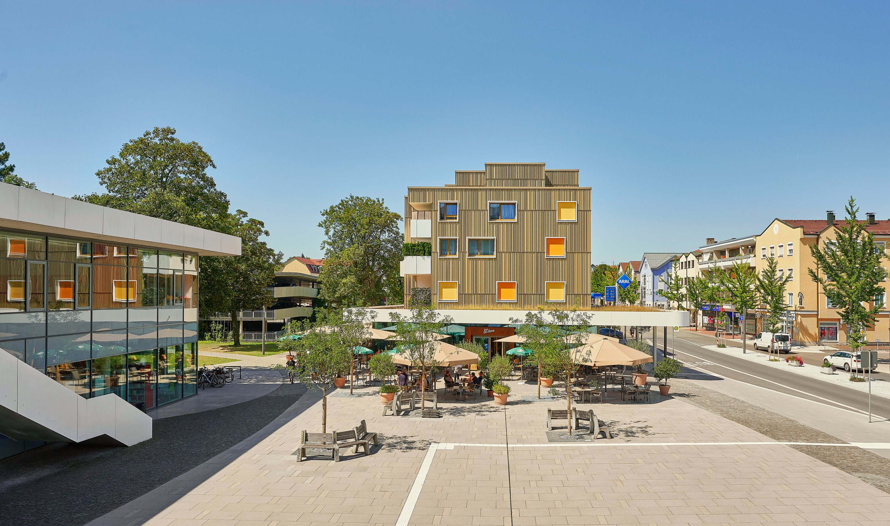 Project by Behnisch Architekturbuero, Mixed-use Building Rathausplatz. Modern building with a wooden facade and colorful shades over a square caf&eacute; with parasols, benches and the surrounding streets and buildings.