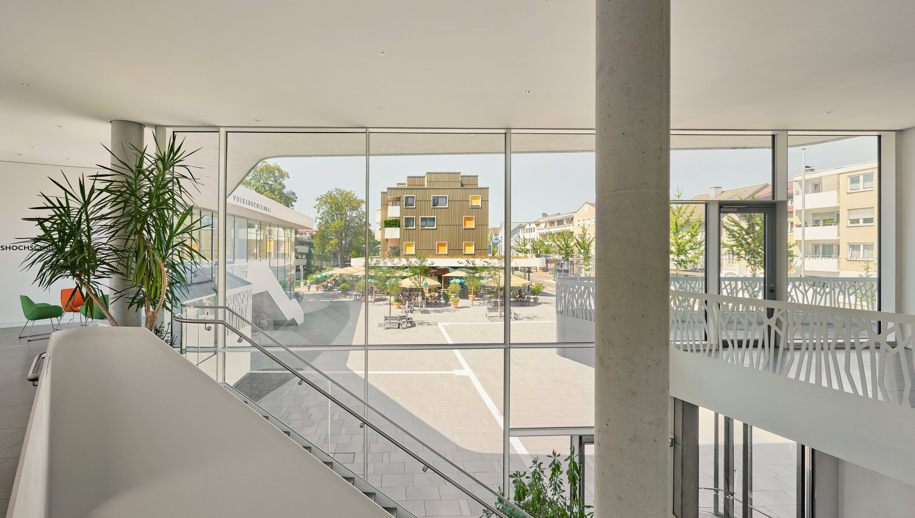 Project by Behnisch Architekturbuero, Mixed-use Building Rathausplatz. Interior view with staircase and large windows overlooking the plaza with caf&eacute; seating and a modern wood-clad building outside.