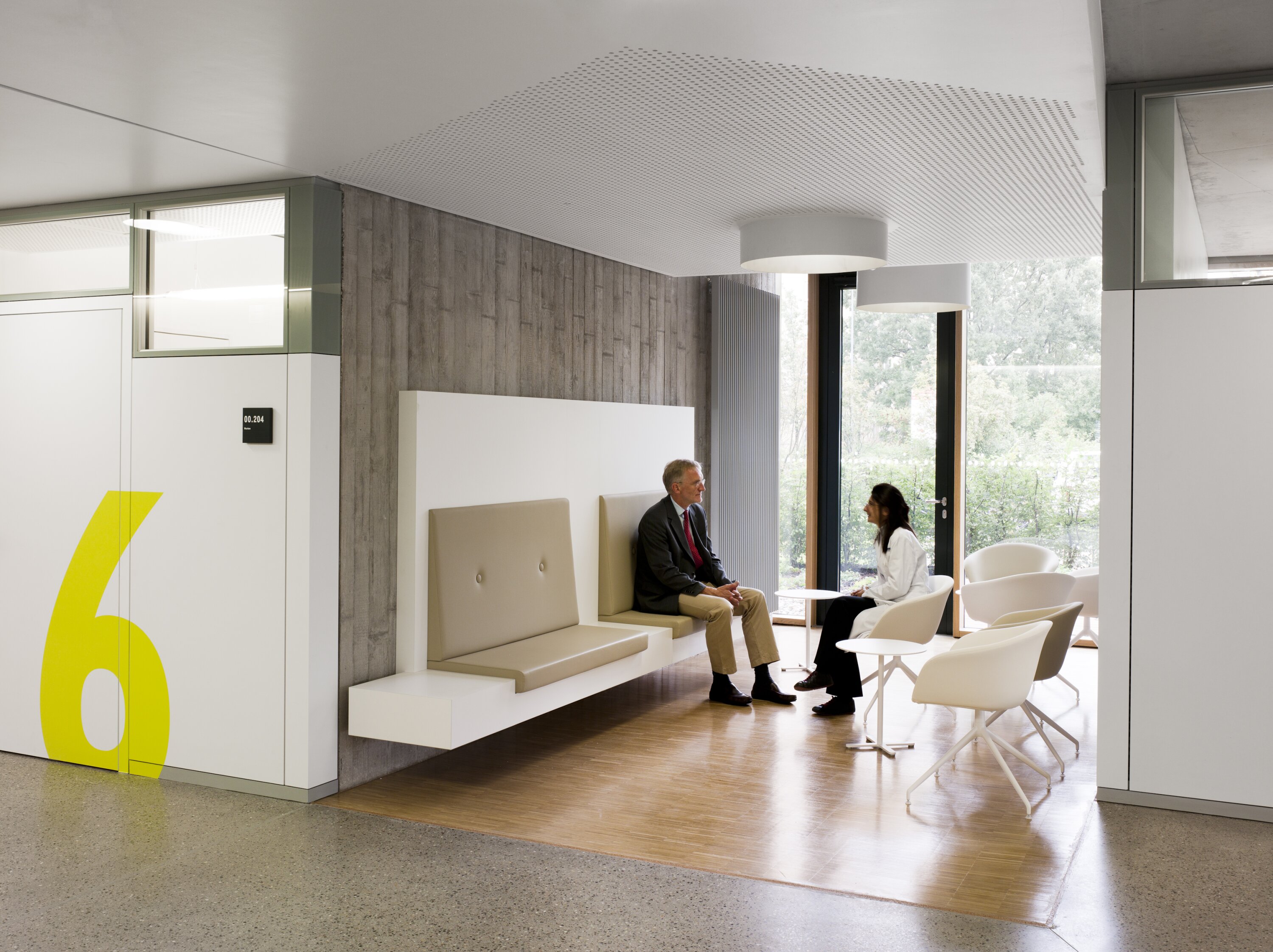 Project by Behnisch Architekturbuero, NCT National Center for Tumour Diseases. Two people are sitting and talking in the lounge area with built-in benches, white chairs, large windows and a yellow 6 on the wall.