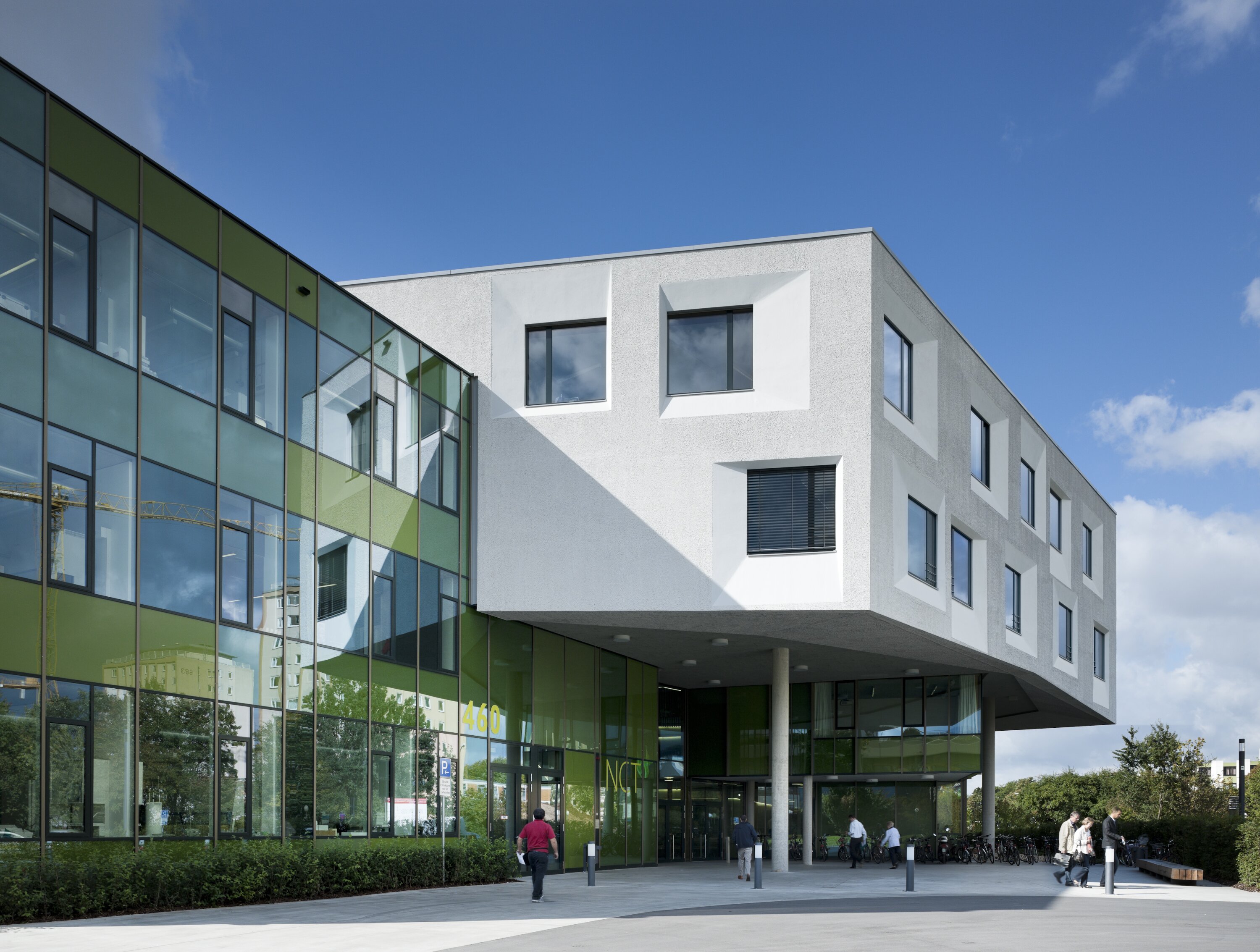 Project by Behnisch Architekturbuero, NCT National Center for Tumour Diseases. Modern building with white cantilevered upper floors and green glass facade, built on pillars, with people walking near the entrance under a blue sky.