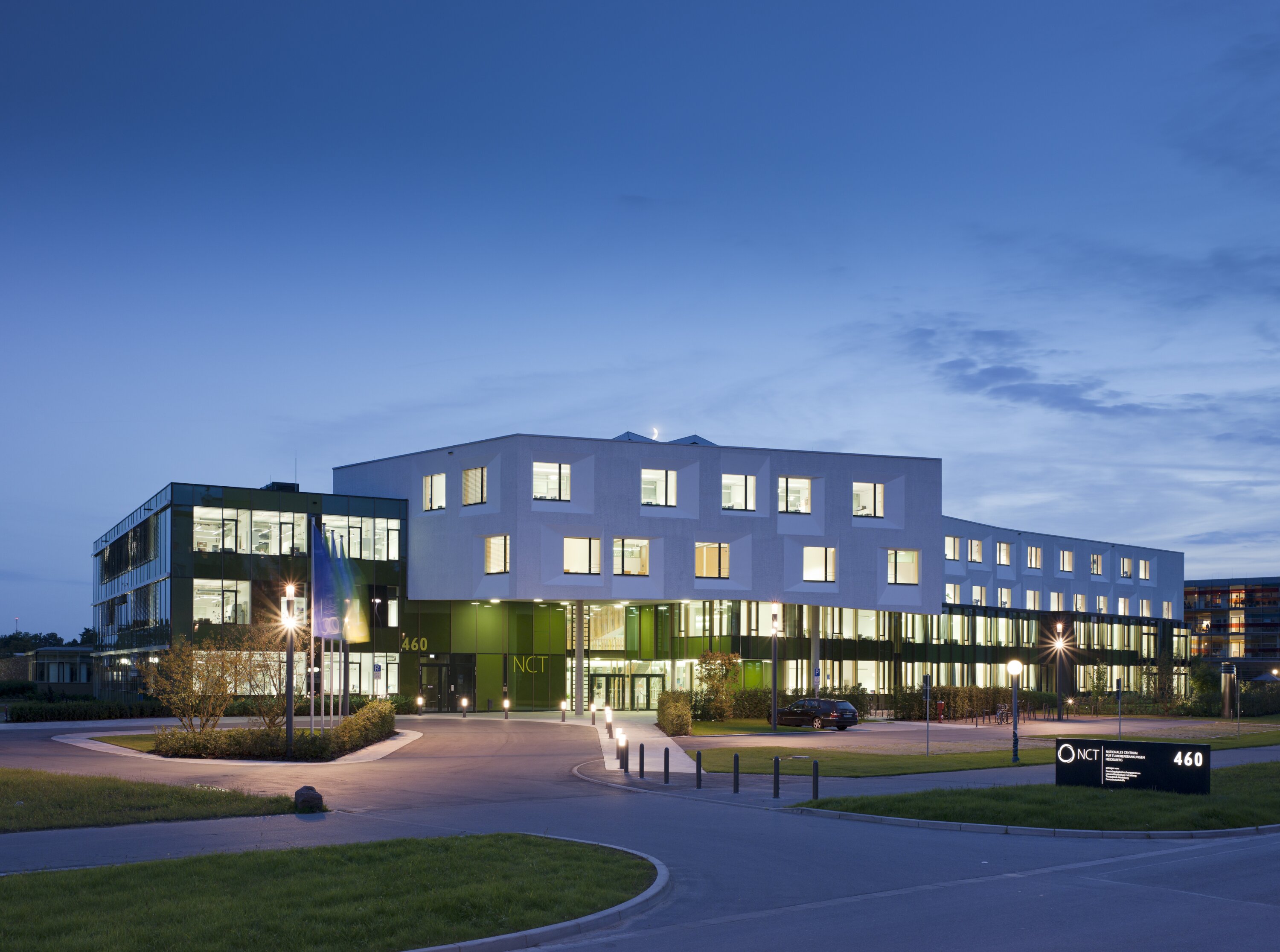 Project by Behnisch Architekturbuero, NCT National Center for Tumour Diseases. Modern building at dusk with illuminated windows, green glass facade and an entrance surrounded by paths, grass and streetlights.