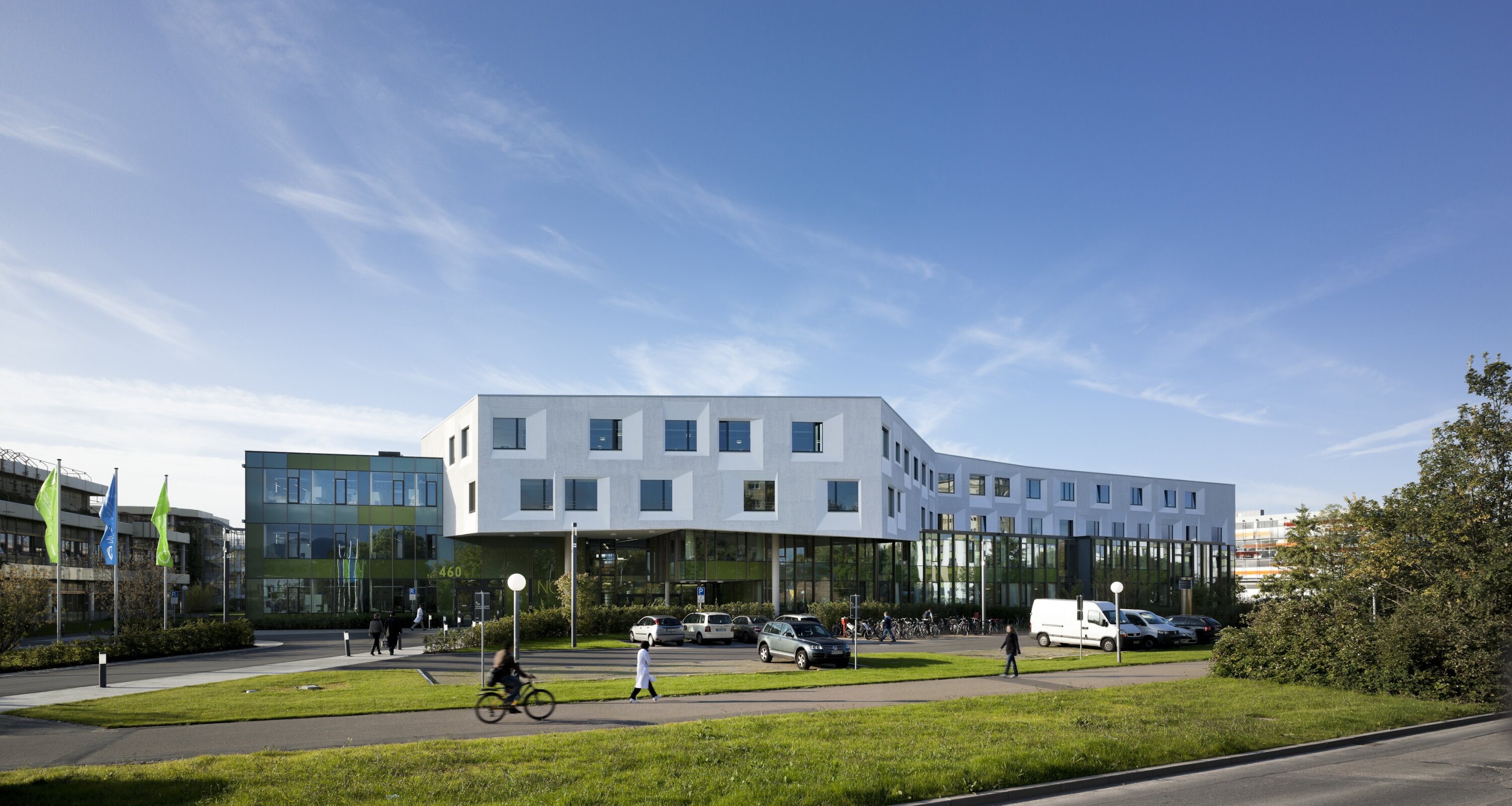 Project by Behnisch Architekturbuero, NCT National Center for Tumour Diseases. Modern building with white and glass facades, surrounded by grass, parked cars, and pedestrians under a blue sky.
