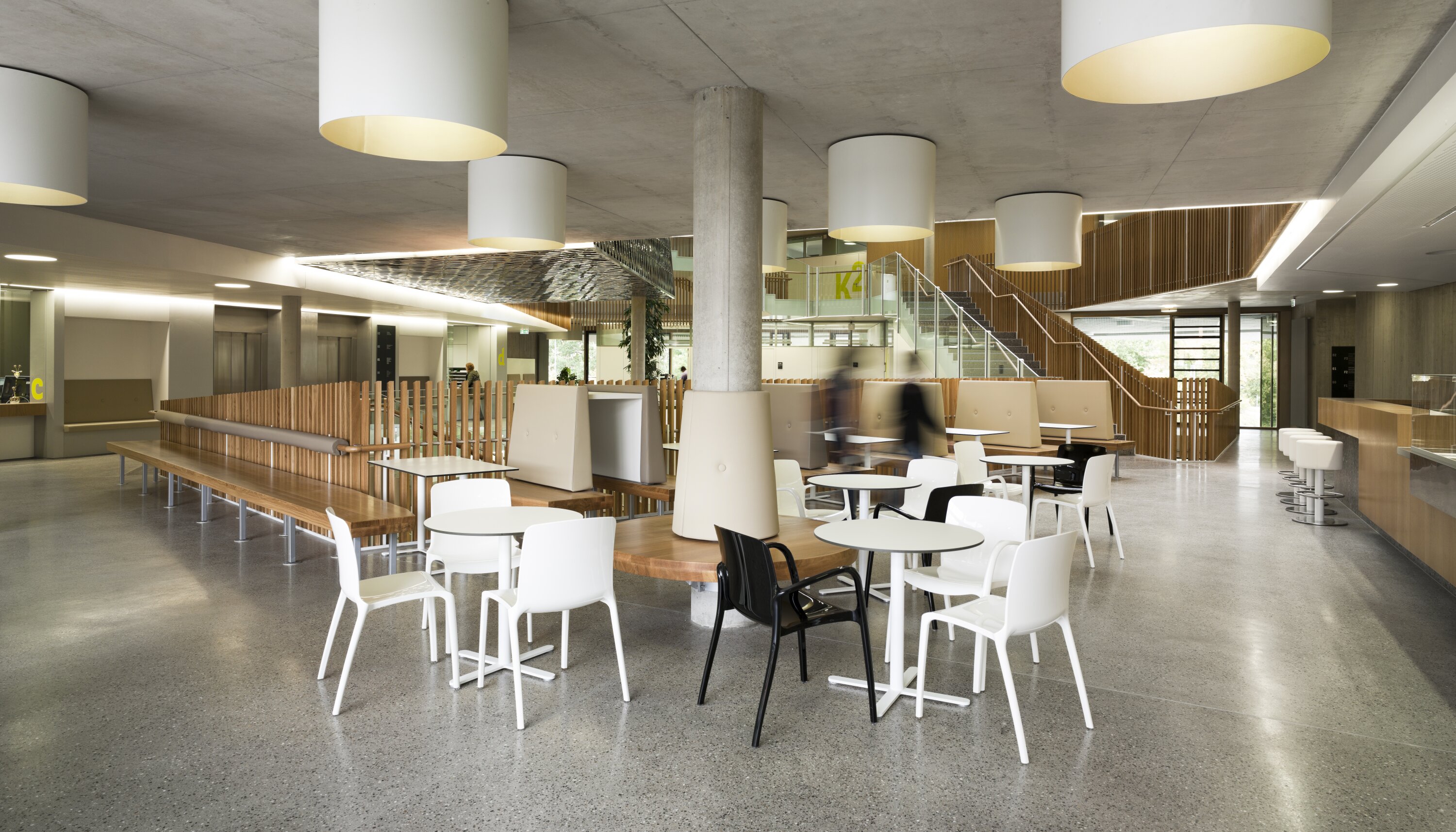 Project by Behnisch Architekturbuero, NCT National Center for Tumour Diseases. Open cafeteria with tables and chairs, wooden benches, large ceiling lights, stairs and people moving around in the modern common room.