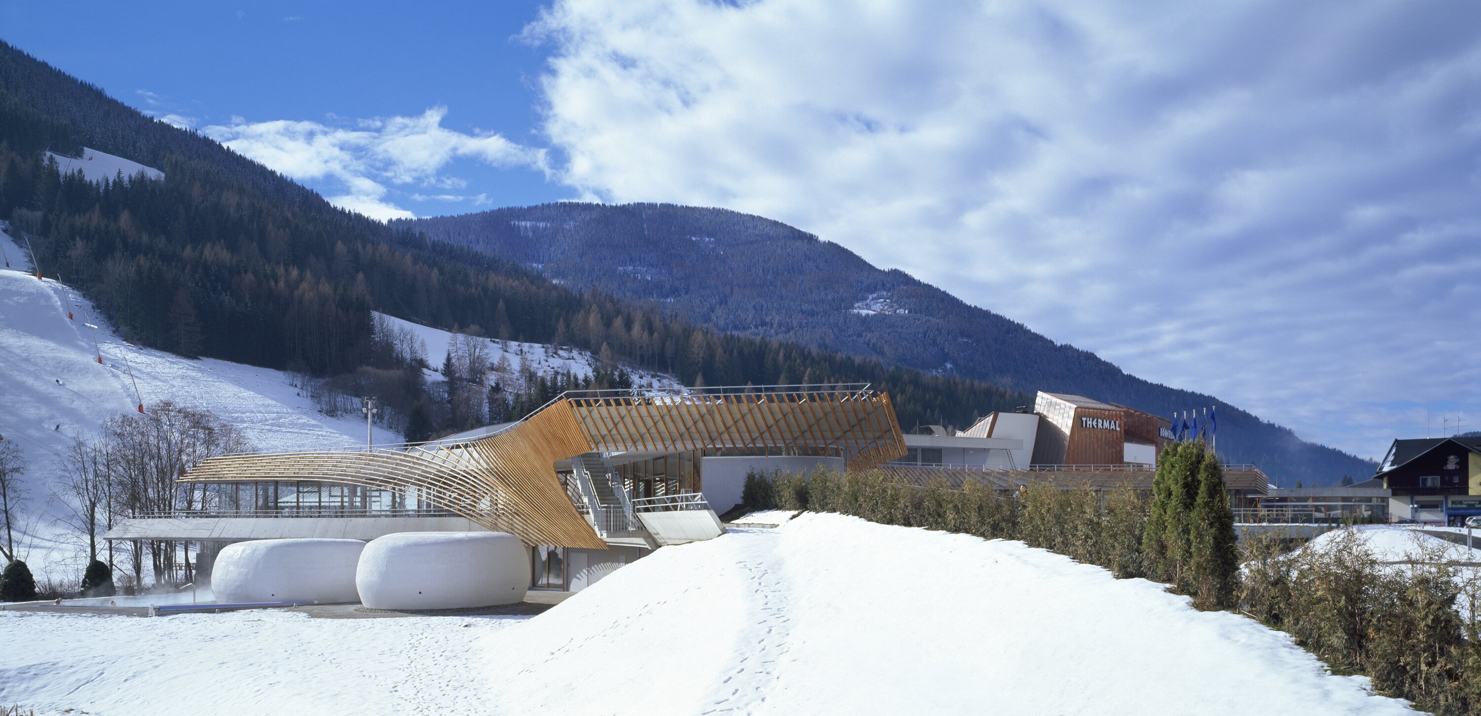 Project by Behnisch Architekturbuero, Thermal Roemerbad. Modern spa complex with curved wooden structure and domed buildings in a snowy mountain landscape under a cloudy blue sky.