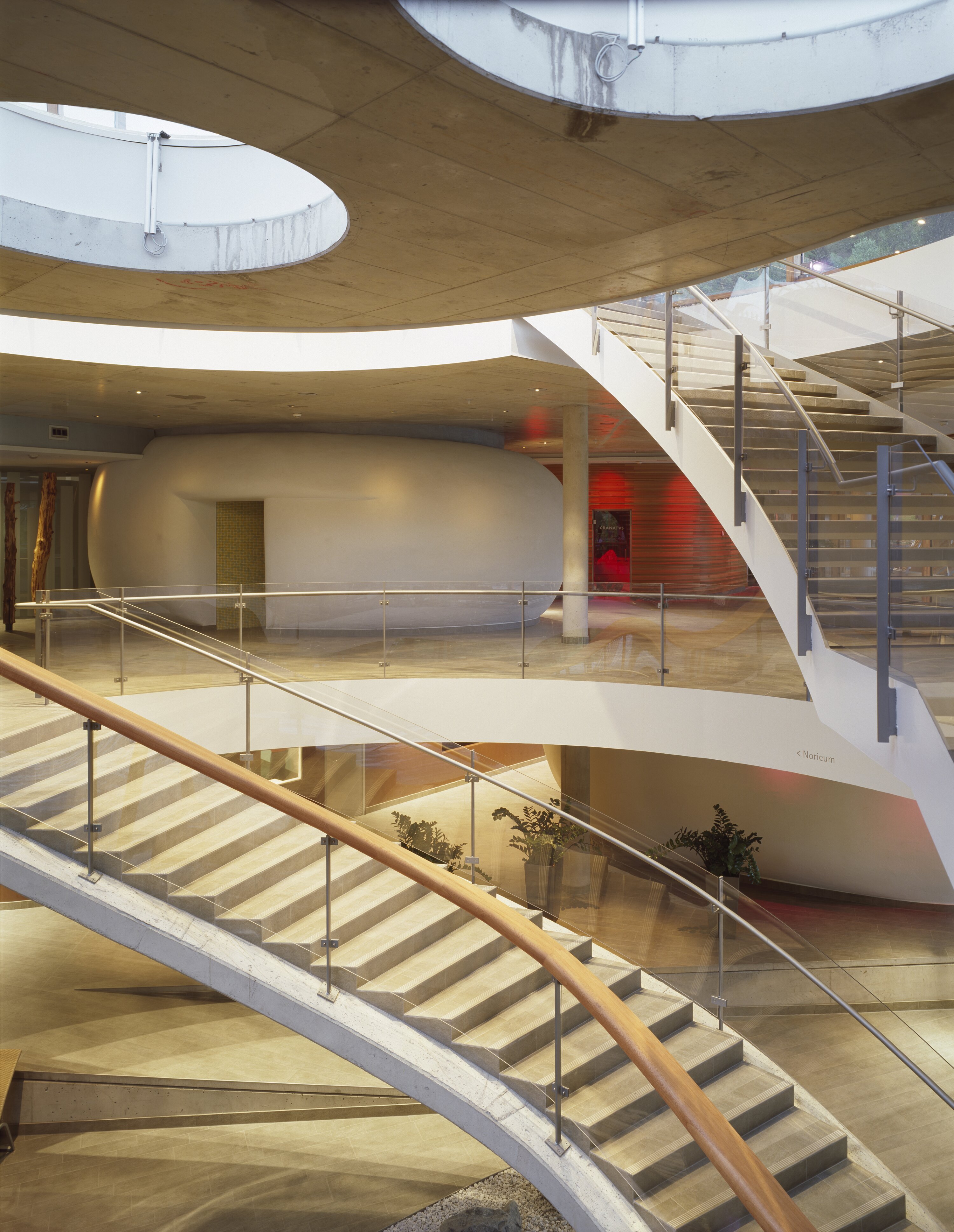 Project by Behnisch Architekturbuero, Thermal Roemerbad. Multi-level interior with curved staircases, glass railings, circular skylights, and smooth white walls with warm accent lighting.