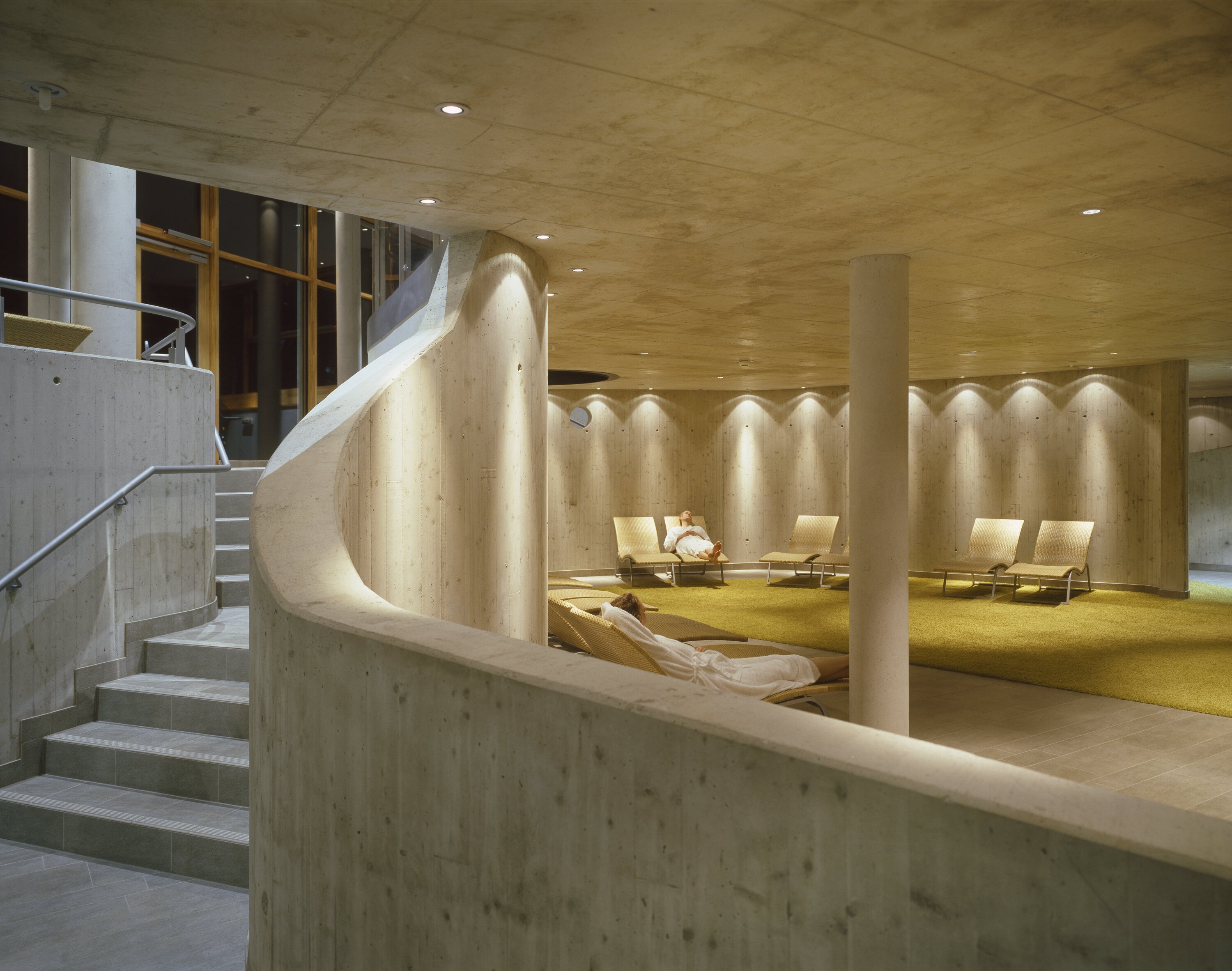 Project by Behnisch Architekturbuero, Thermal Roemerbad. Curved concrete interior with stair, soft lighting, and lounge chairs where people relax in a quiet spa area.