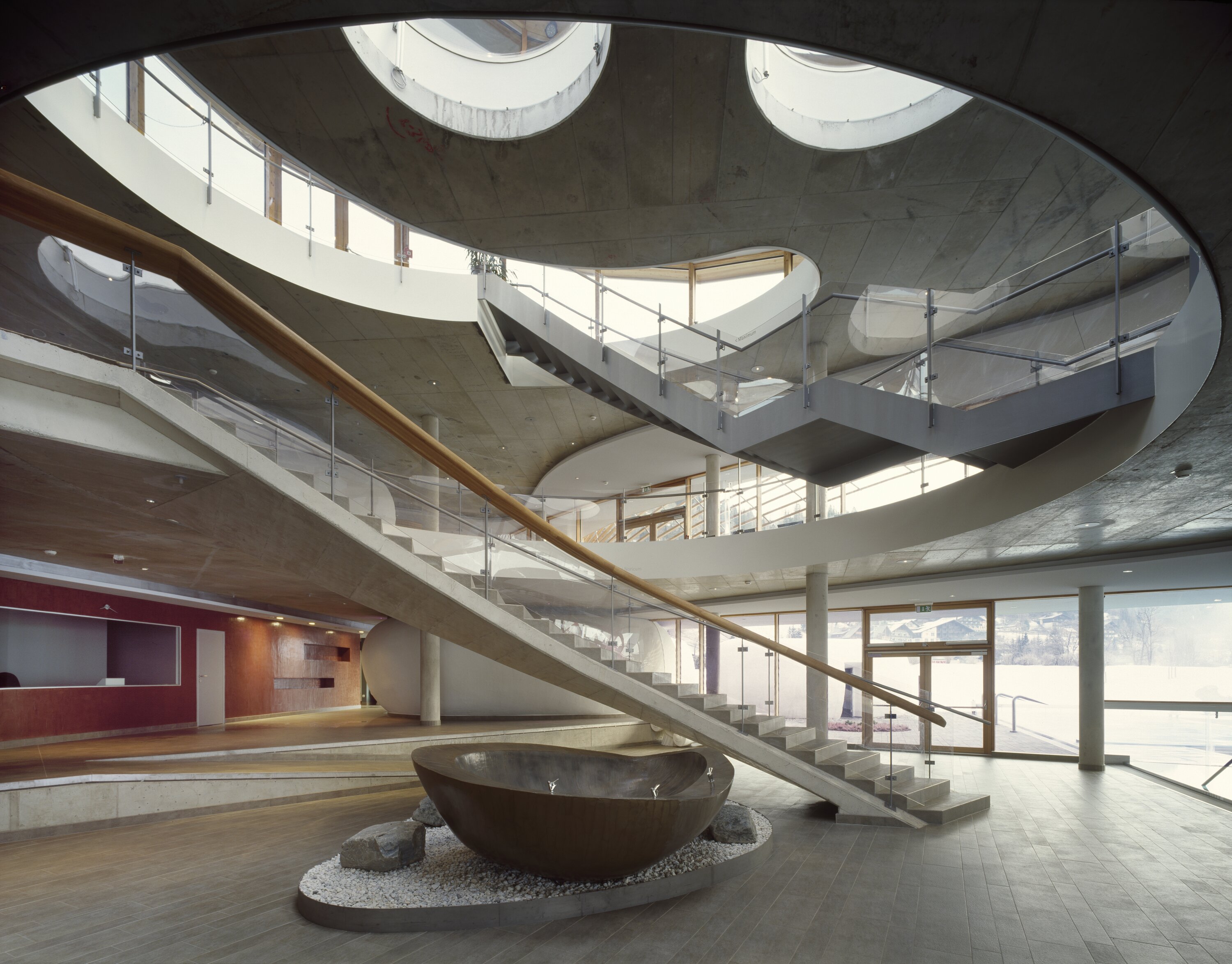 Project by Behnisch Architekturbuero, Thermal Roemerbad. Curved interior atrium with sweeping staircase, circular skylights, glass railings, and a large bowl-shaped fountain at center.