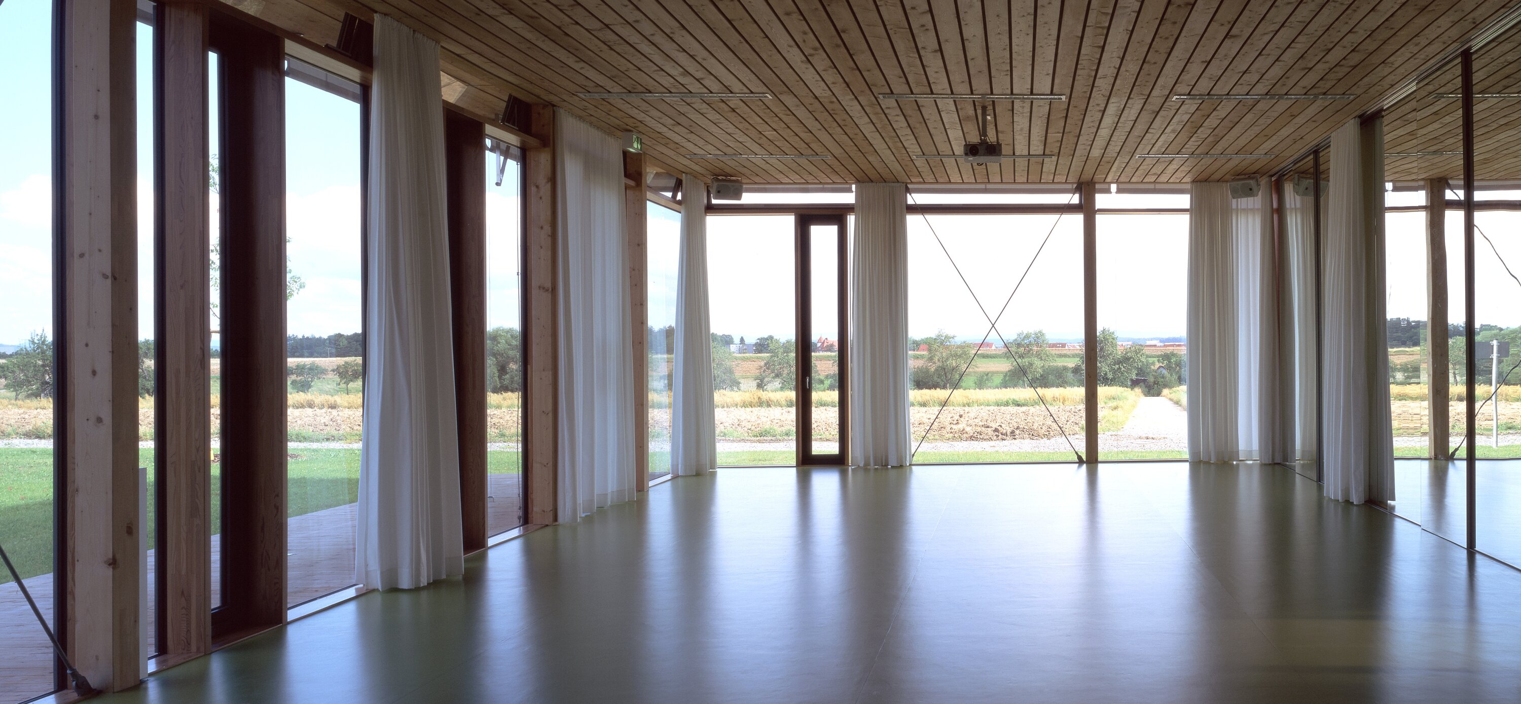 Project by Behnisch Architekturbuero, Hilde Domin School. Empty room with wooden ceiling, green floor, and floor-to-ceiling windows with curtains overlooking fields and trees.