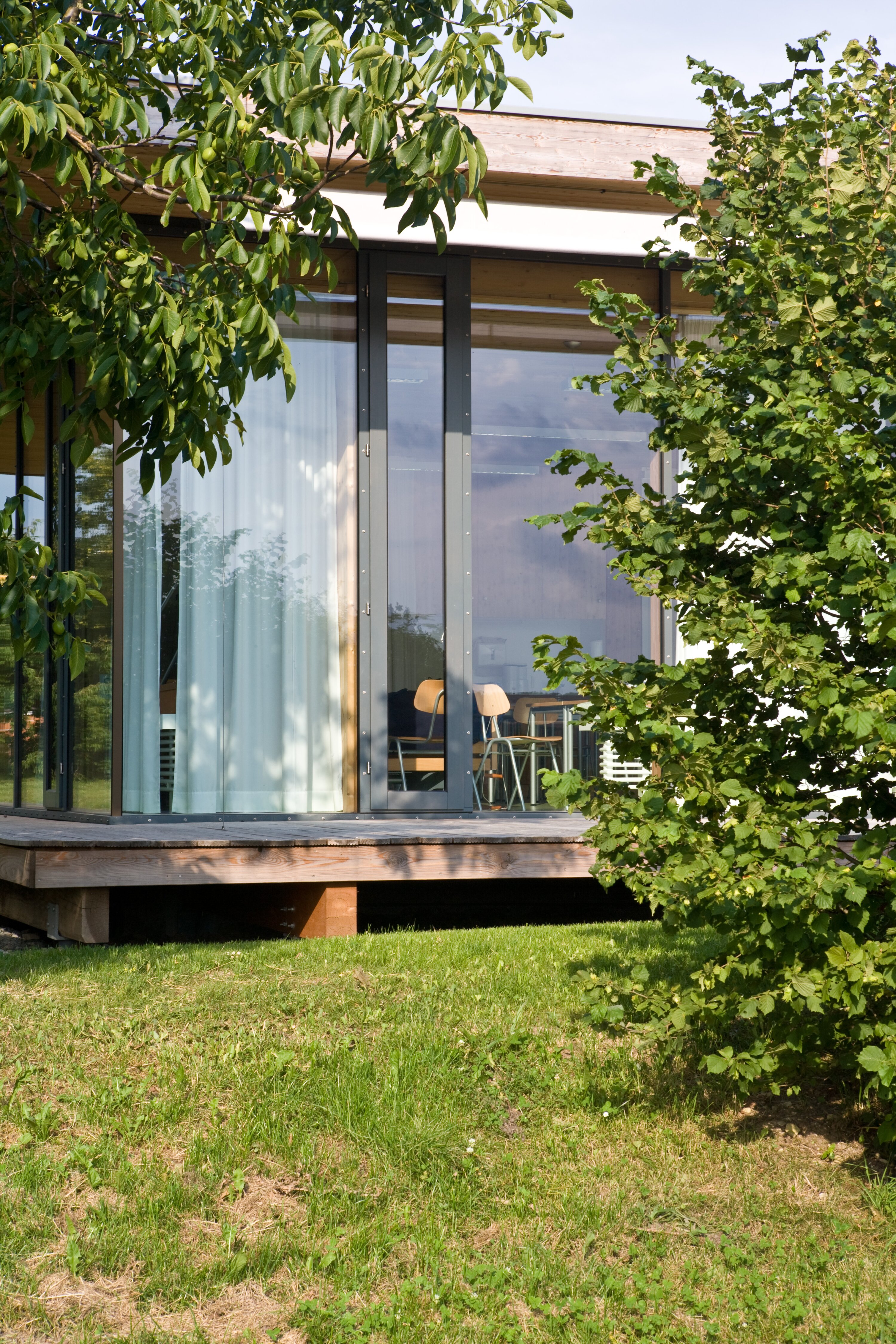 Project by Behnisch Architekturbuero, Hilde Domin School. Exterior view of the building with a floor-to-ceiling glass facade that can be shaded with curtains. The elevated building is visible through leafy trees and sits on a lawn with a wooden terrace.