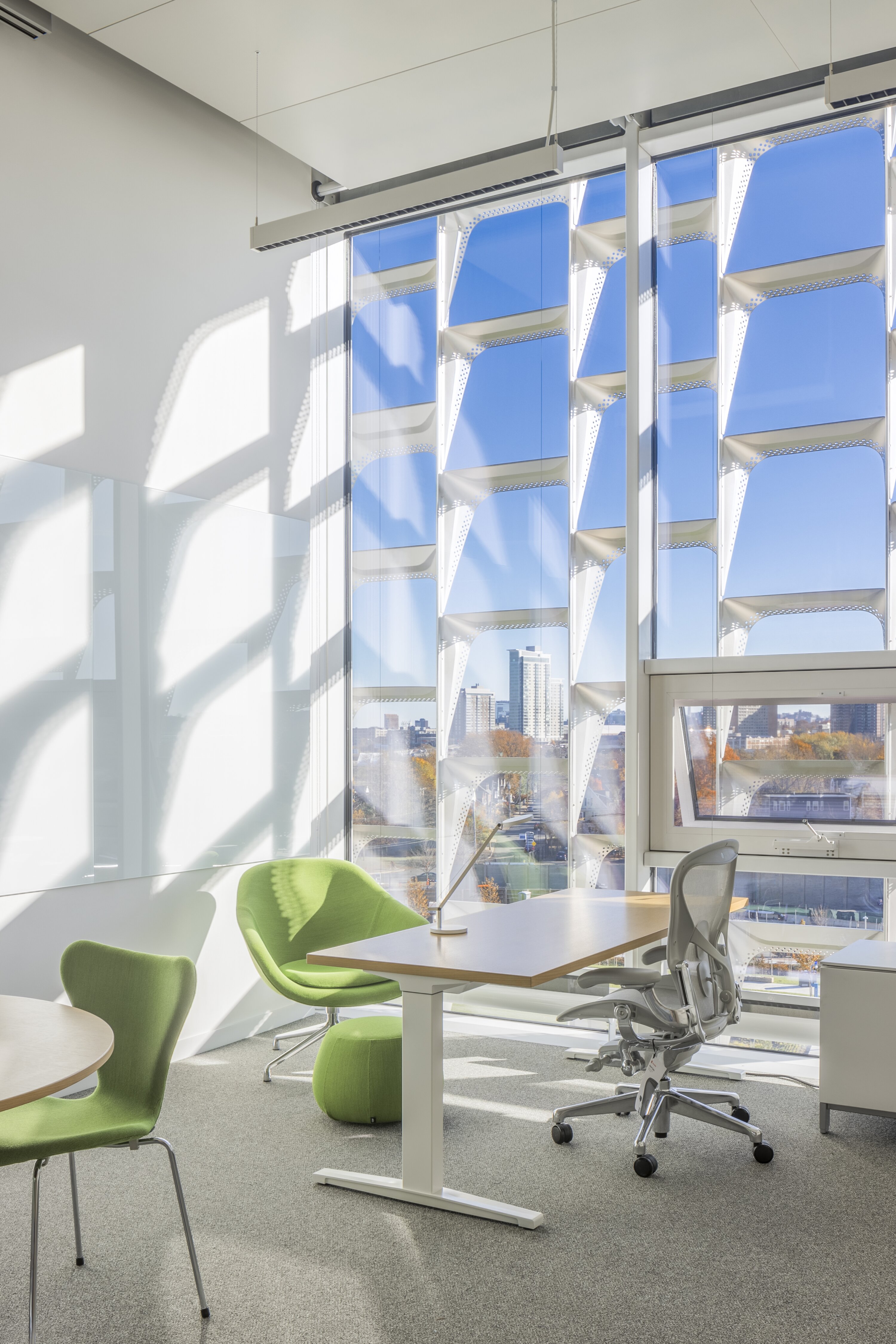 Project by Behnisch Architekturbuero, Harvard University Science and Engineering Complex (SEC). A small office corner with a desk, chair, and seating made of green plants by a large window. Through the window, you can see the sunshade panels and the city.
