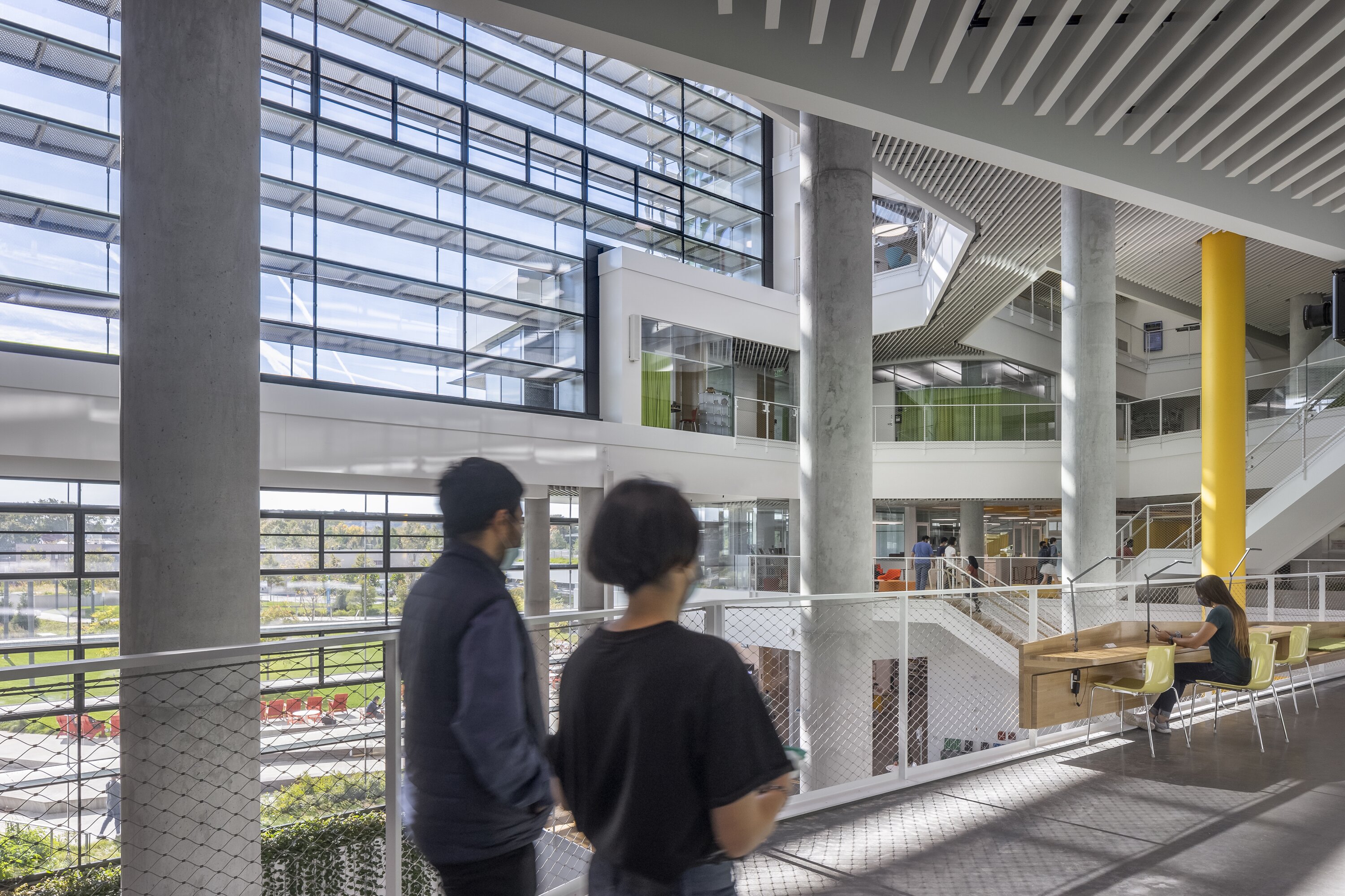Project by Behnisch Architekturbuero, Harvard University Science and Engineering Complex (SEC). People walking along an upper-level walkway in a bright atrium with large windows, concrete columns, and a yellow column, with seating nearby where student are working.
