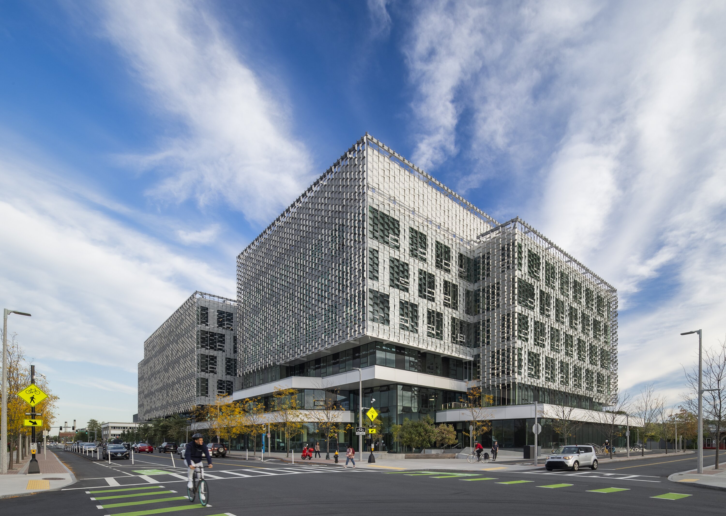 Project by Behnisch Architekturbuero, Harvard University Science and Engineering Complex (SEC). Full view of the multi-story building with the sunshade panels. The building is located on a street corner. Cyclists, pedestrians, and cars are passing by.