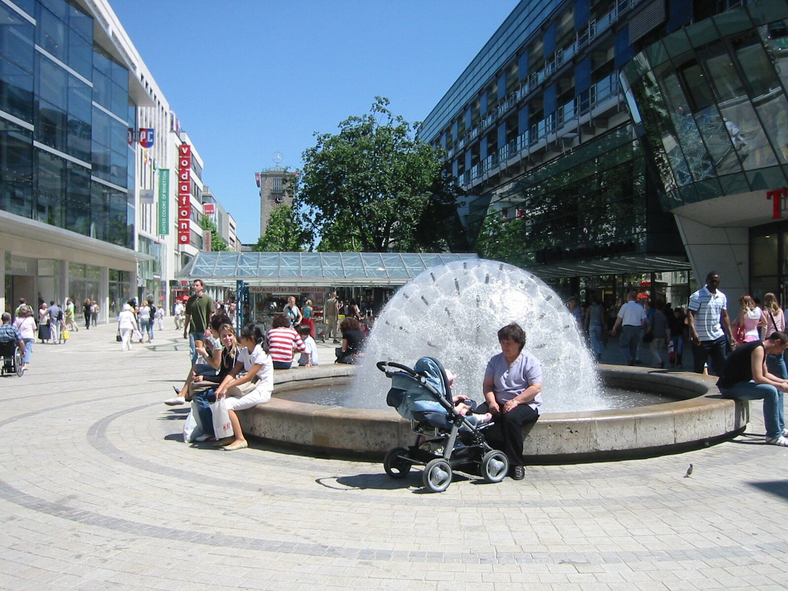 Project by Behnisch Architekturbuero, Refurbishment and Modernization K&ouml;nigsstra&szlig;e. People sit around a circular fountain in a busy pedestrian plaza with shops and modern buildings under a clear sky.