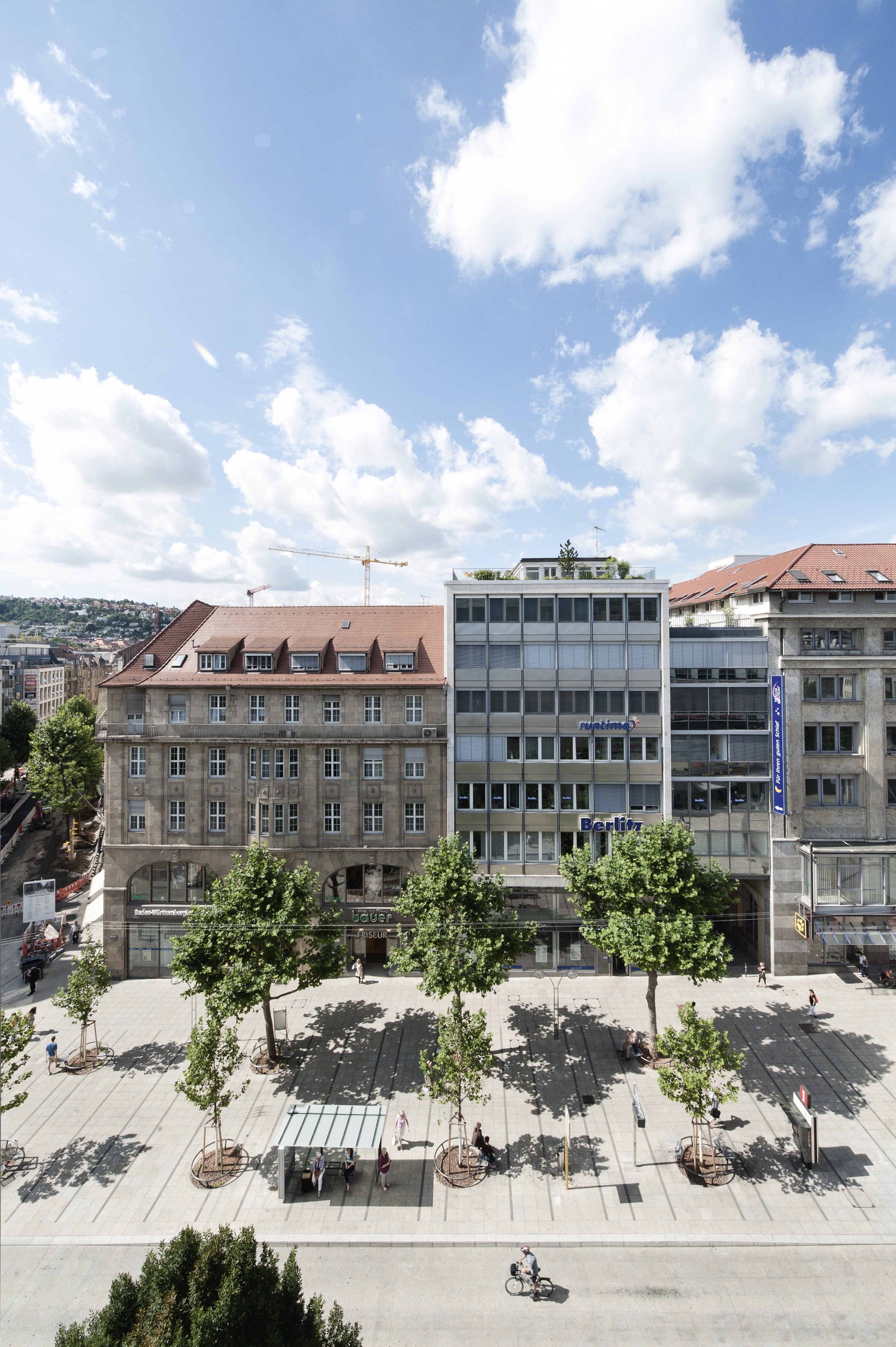 Project by Behnisch Architekturbuero, Refurbishment and Modernization K&ouml;nigsstra&szlig;e. Row of trees along a paved plaza in front of mid-rise buildings. Pedestrians are walking by.