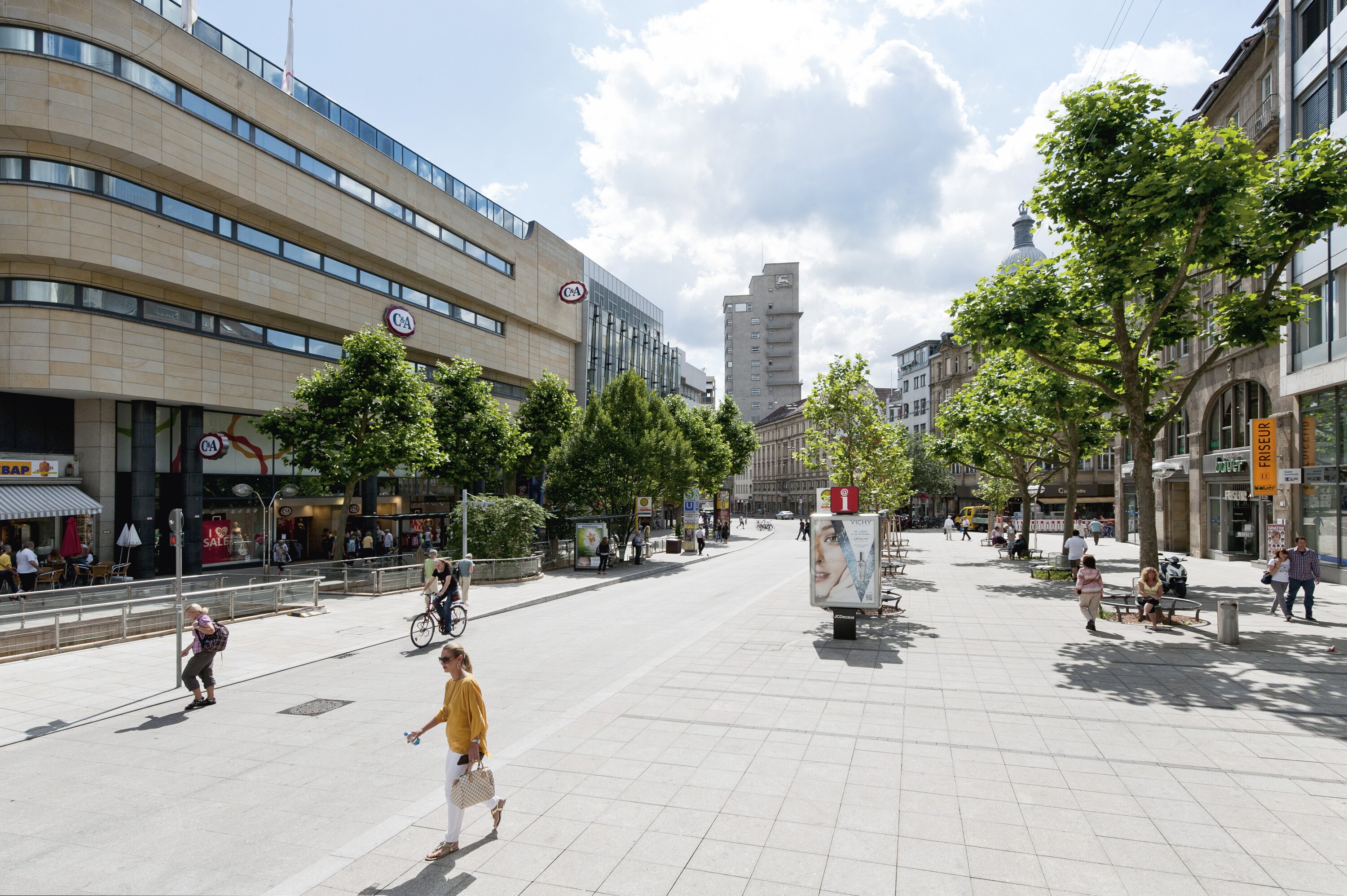 Project by Behnisch Architekturbuero, Refurbishment and Modernization K&ouml;nigsstra&szlig;e. Pedestrian street with trees, shops and cafes. People are walking and cycling on the road. Benches along a wide paved urban plaza under a cloudy sky.