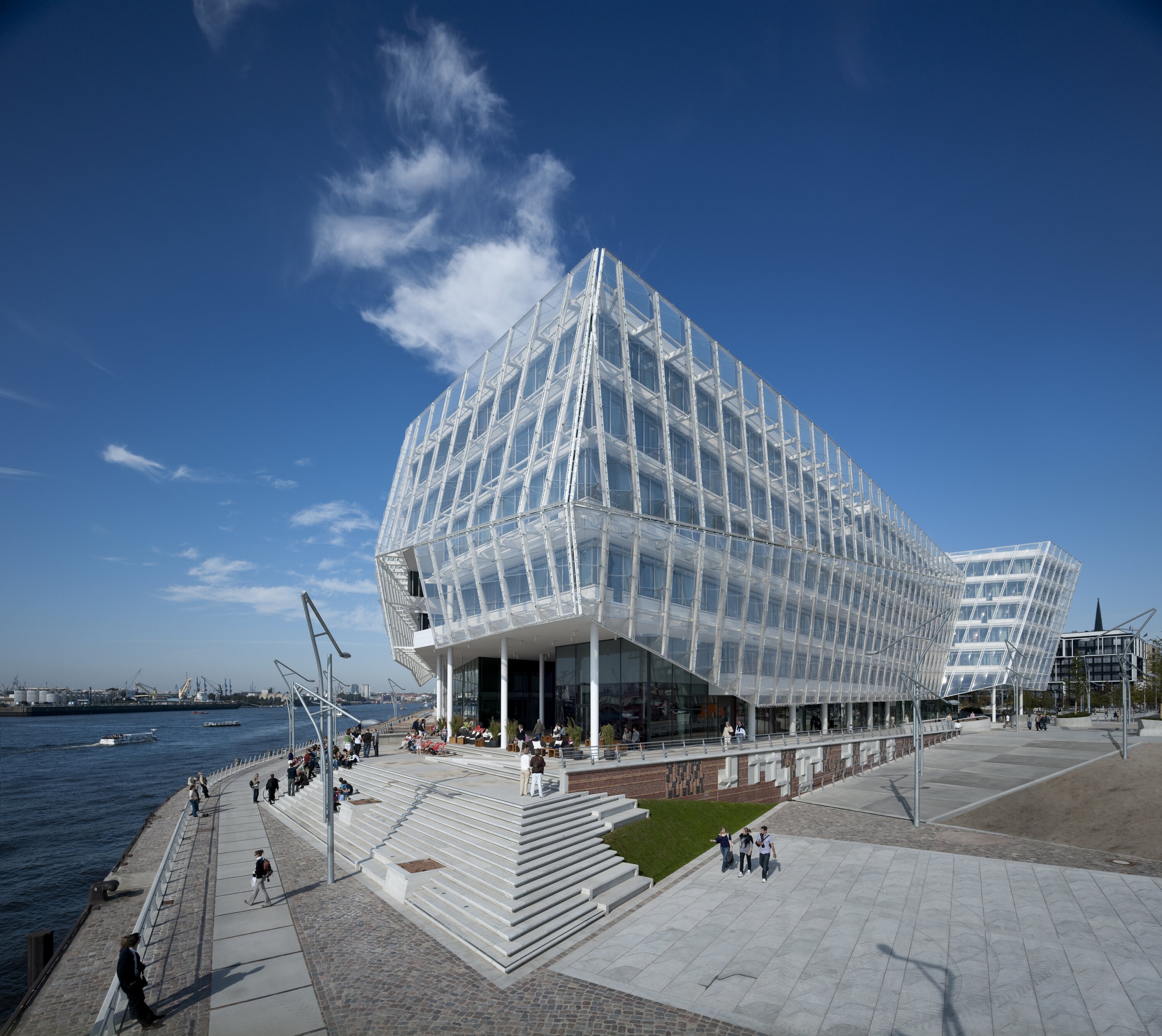 Project by Behnisch Architekturbuero, Unilever Headquarters for Germany, Austria, and Switzerland. The building is enveloped in an ETFE membrane that protects the primary building envelope. The angular facade sits on the banks of the Northelbe River in Hamburg, with people on the steps and promenade beneath a blue sky dotted with clouds.