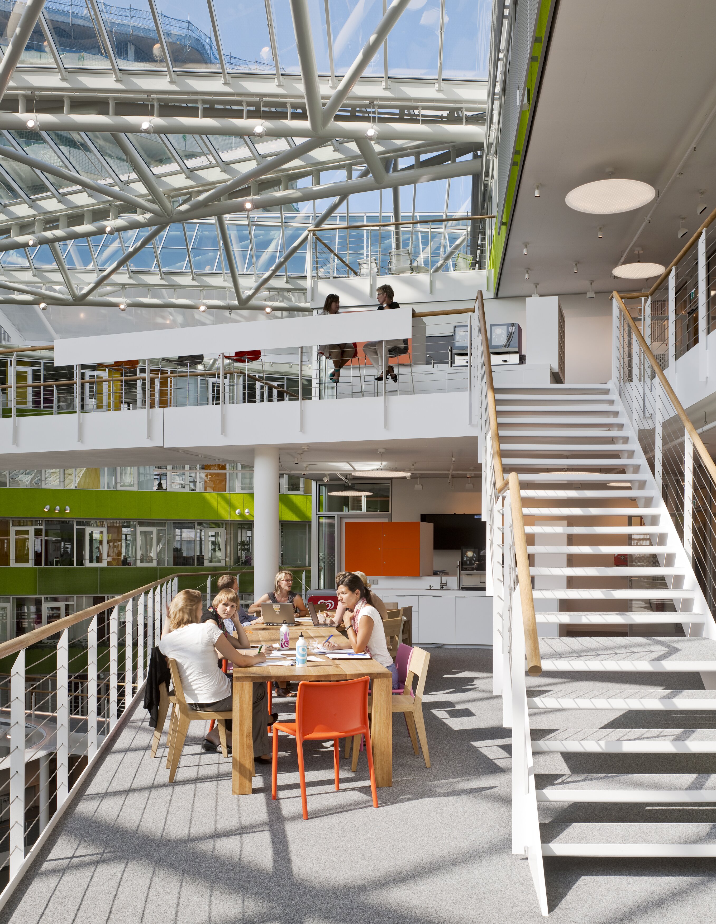 Project by Behnisch Architekturbuero, Unilever Headquarters for Germany, Austria, and Switzerland. People sitting at a table and others on an upper walkway in the bright atrium with stairs, glass roof, and green-accented interior spaces.