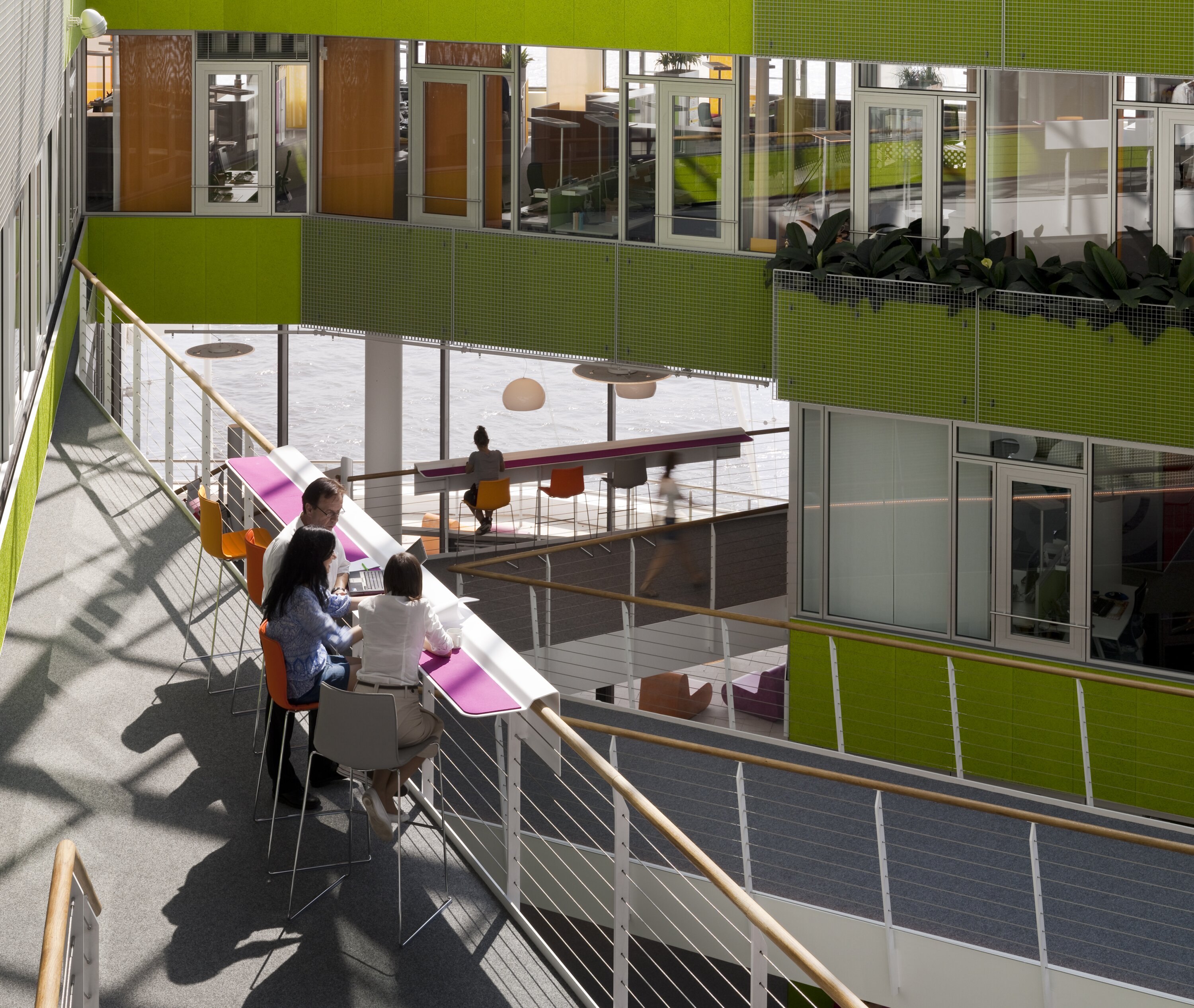 Project by Behnisch Architekturbuero, Unilever Headquarters for Germany, Austria, and Switzerland. People seated at a counter on an upper walkway in a bright atrium with green walls, glass offices, railings, and open views to lower floors.