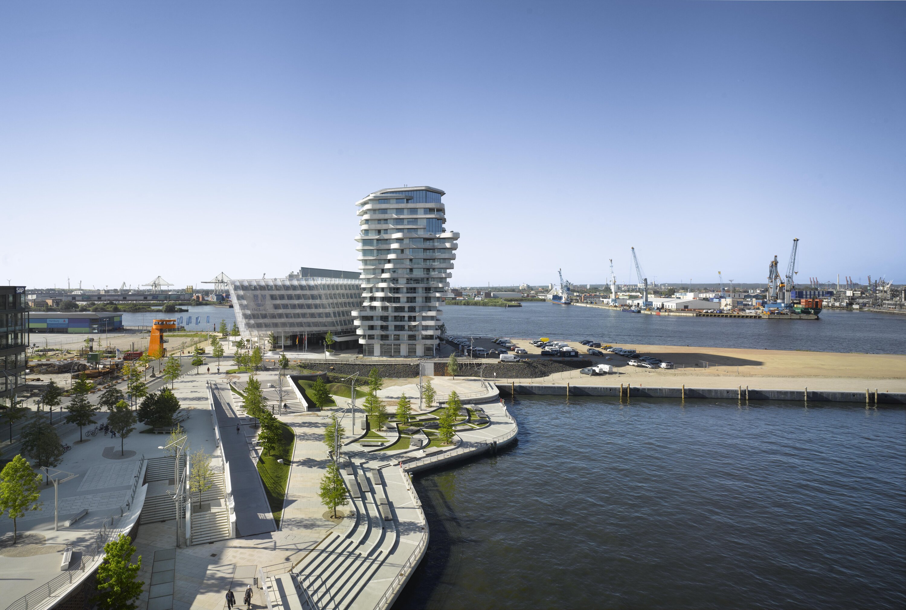 Project by Behnisch Architekturbuero, Marco Polo Tower. Waterfront cityscape with a modern high-rise tower, riverside promenade, and harbor with cranes and industrial buildings in the background.