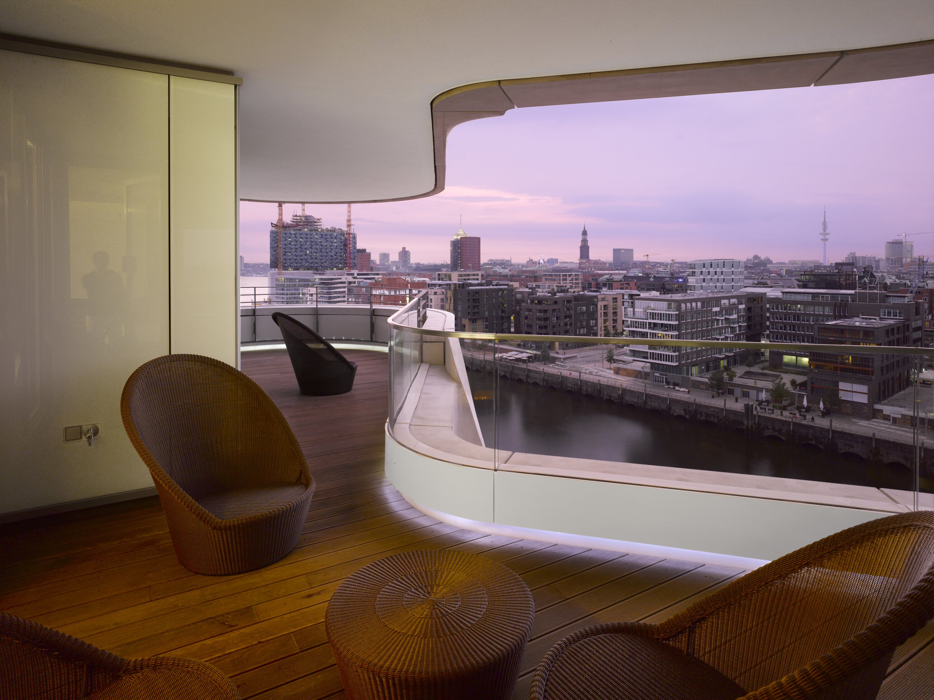 Project by Behnisch Architekturbuero, Marco Polo Tower. Curved balcony with wicker chairs overlooking the river and modern city skyline at dusk, viewed from a furnished interior terrace.