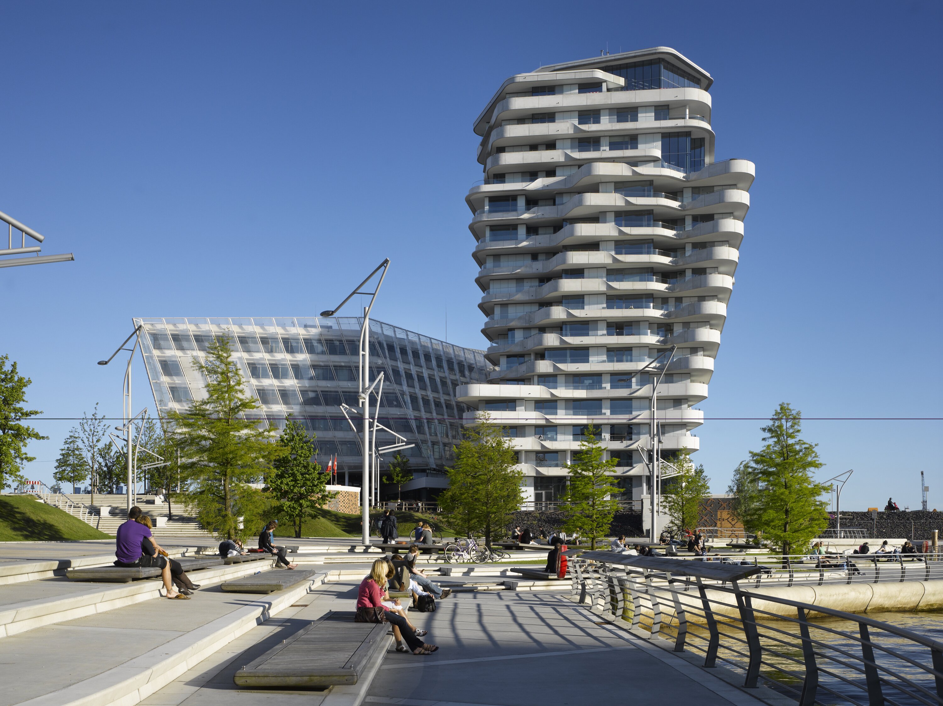 Project by Behnisch Architekturbuero, Marco Polo Tower. Modern waterfront plaza with people sitting on steps, beside the river, and the high-rise building with curved balconies in the background.
