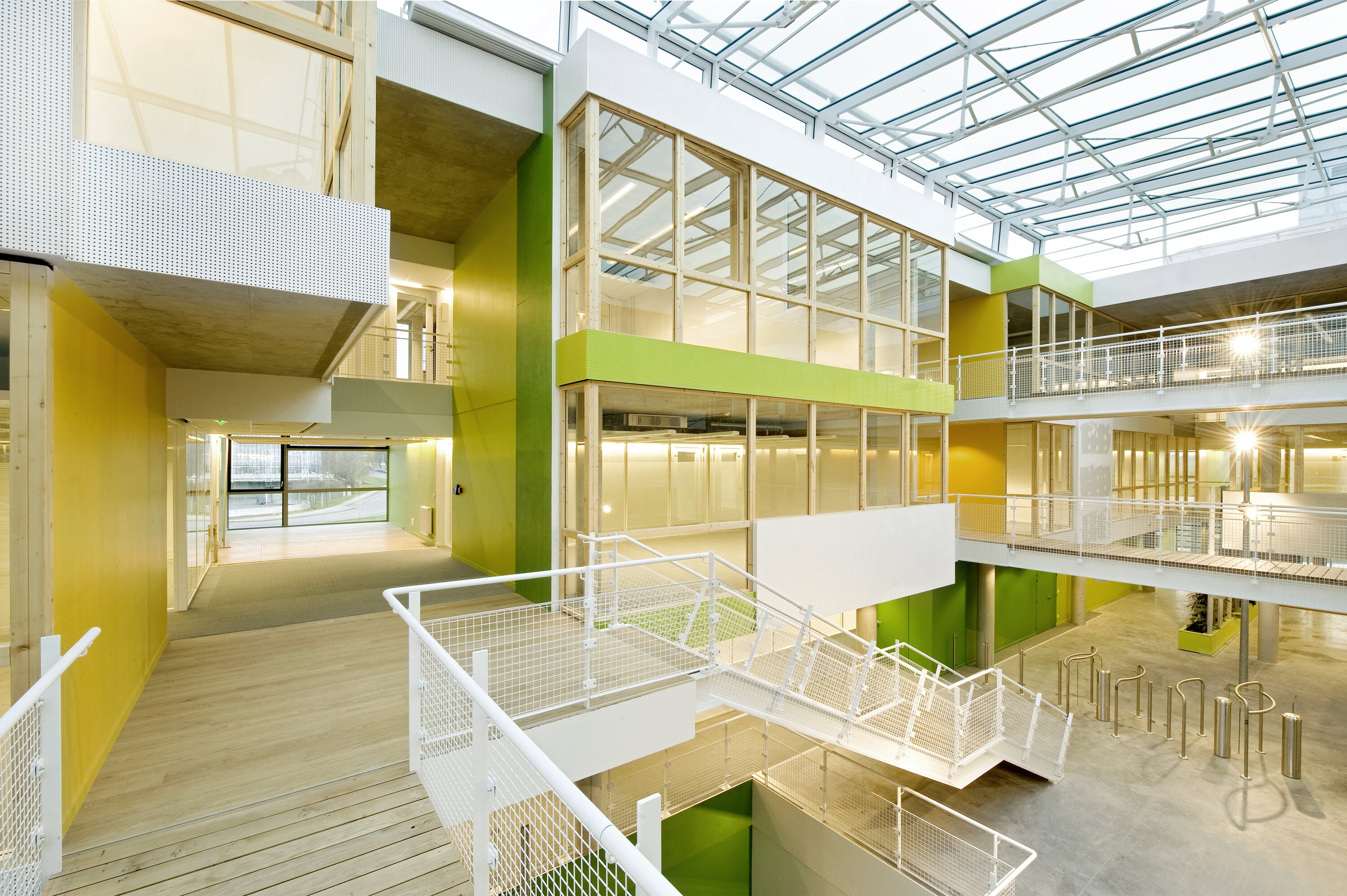 Project by Behnisch Architekturbuero, Digiteo Labs - 3 Research Buildings. Multi-level atrium with green and yellow walls, glass rooms, white railings, and stairs connecting open walkways under a skylit ceiling.