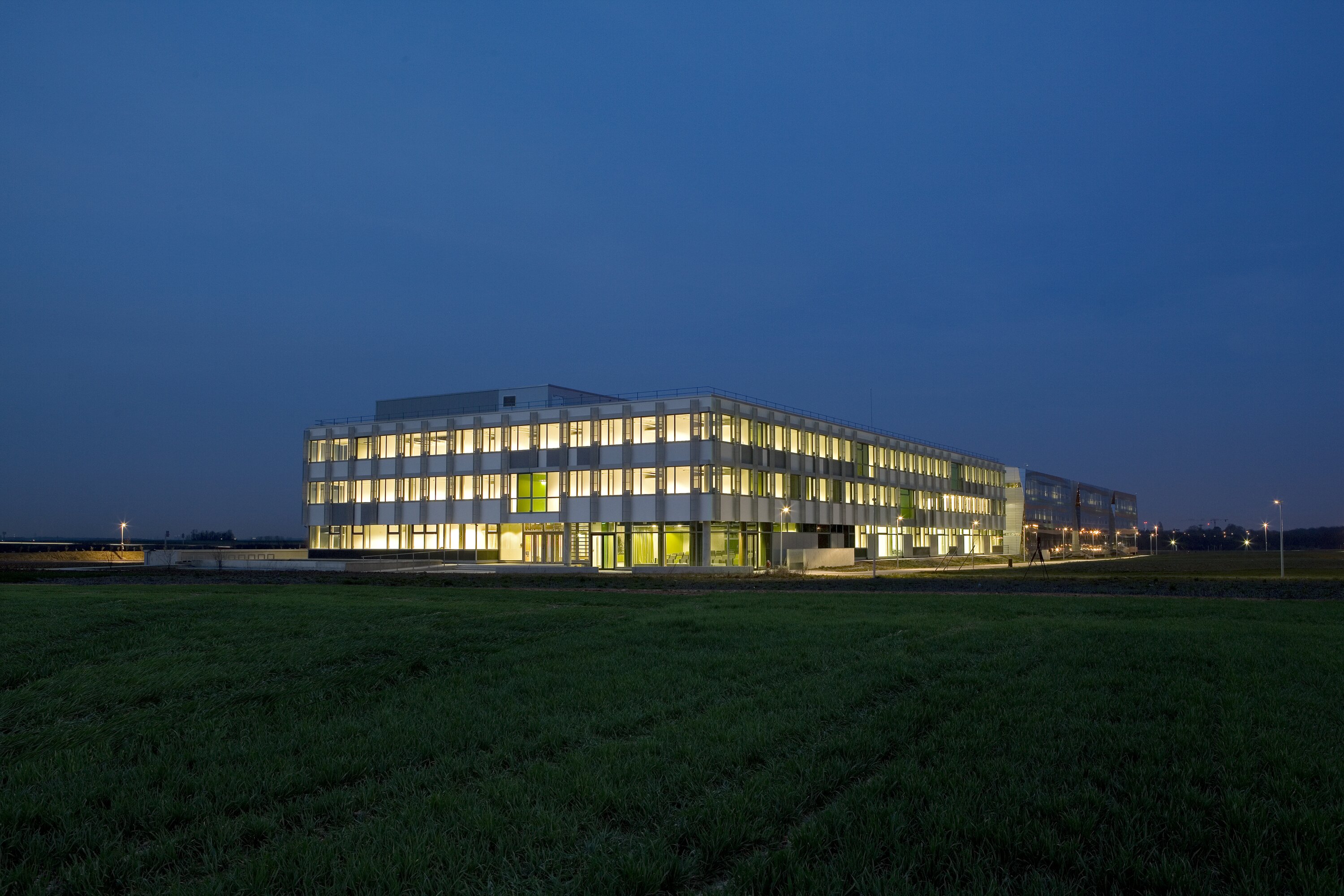 Project by Behnisch Architekturbuero, Digiteo Labs - 3 Research Buildings. Rectangular building with lit windows at dusk, set in an open grassy field under a dark blue sky.