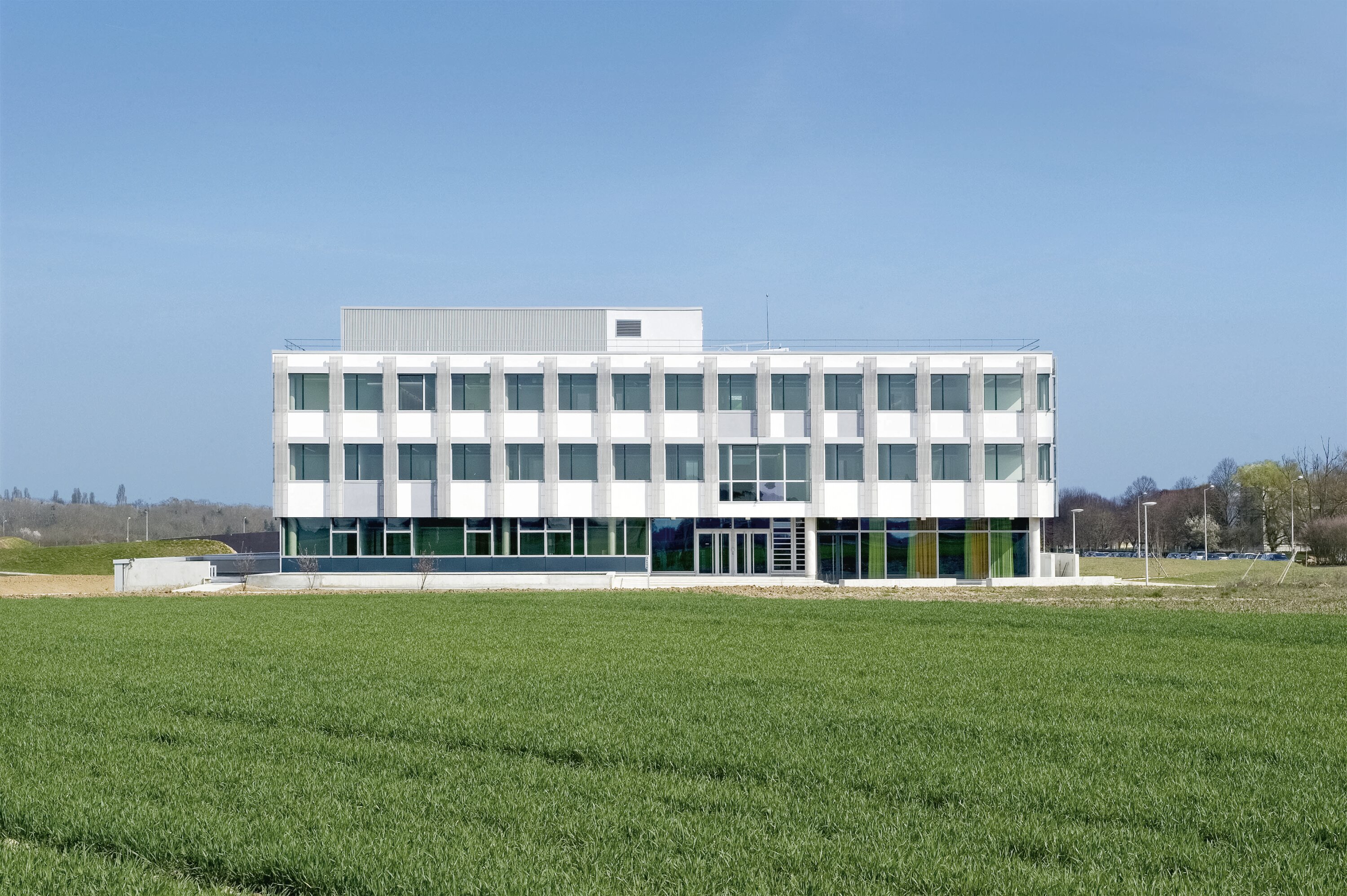 Project by Behnisch Architekturbuero, Digiteo Labs - 3 Research Buildings. The rectangular white office building with grid windows, set behind a wide green field under a clear blue sky.