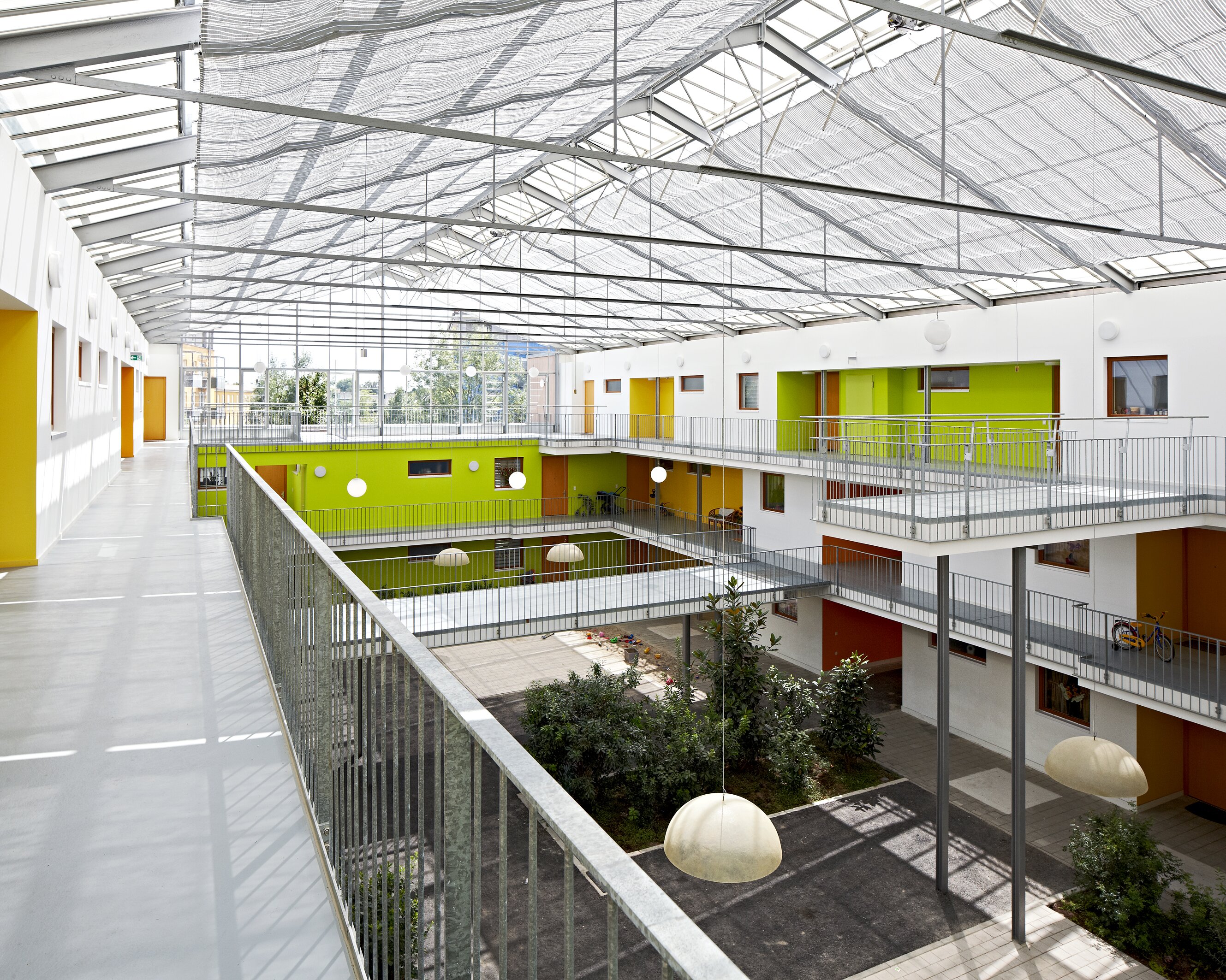 Project by Behnisch Architekturbuero, Residential Building Ingolstadt-Hollerstauden. Interior courtyard of the building with open walkways, green, orange and yellow walls, railings, and a glass roof letting in daylight.