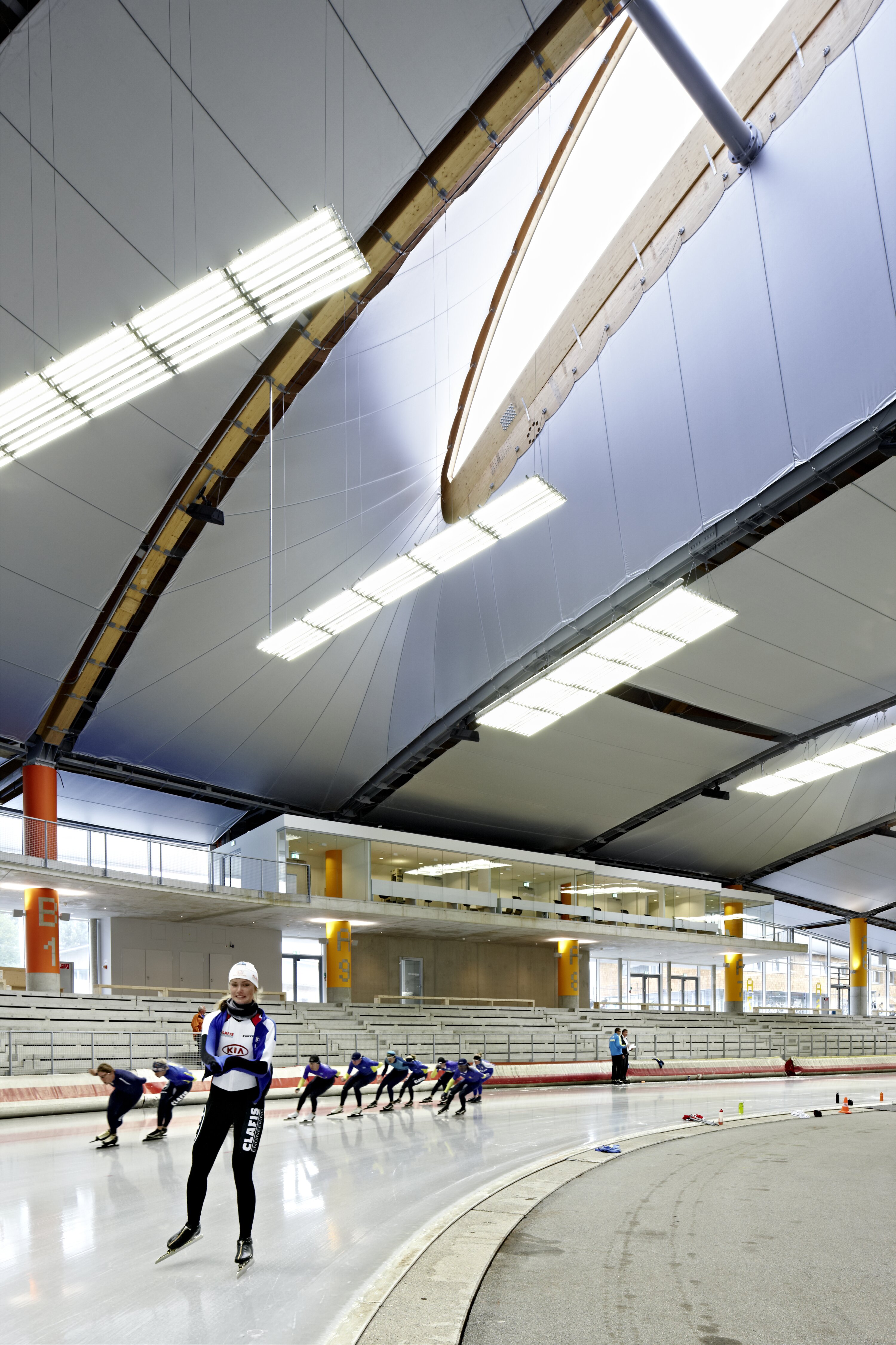 Speed ​​skaters are training in an ice rink. One skater is visible in the foreground, the others are skating a curve. The roof with a large skylight is visible at the top of the picture.