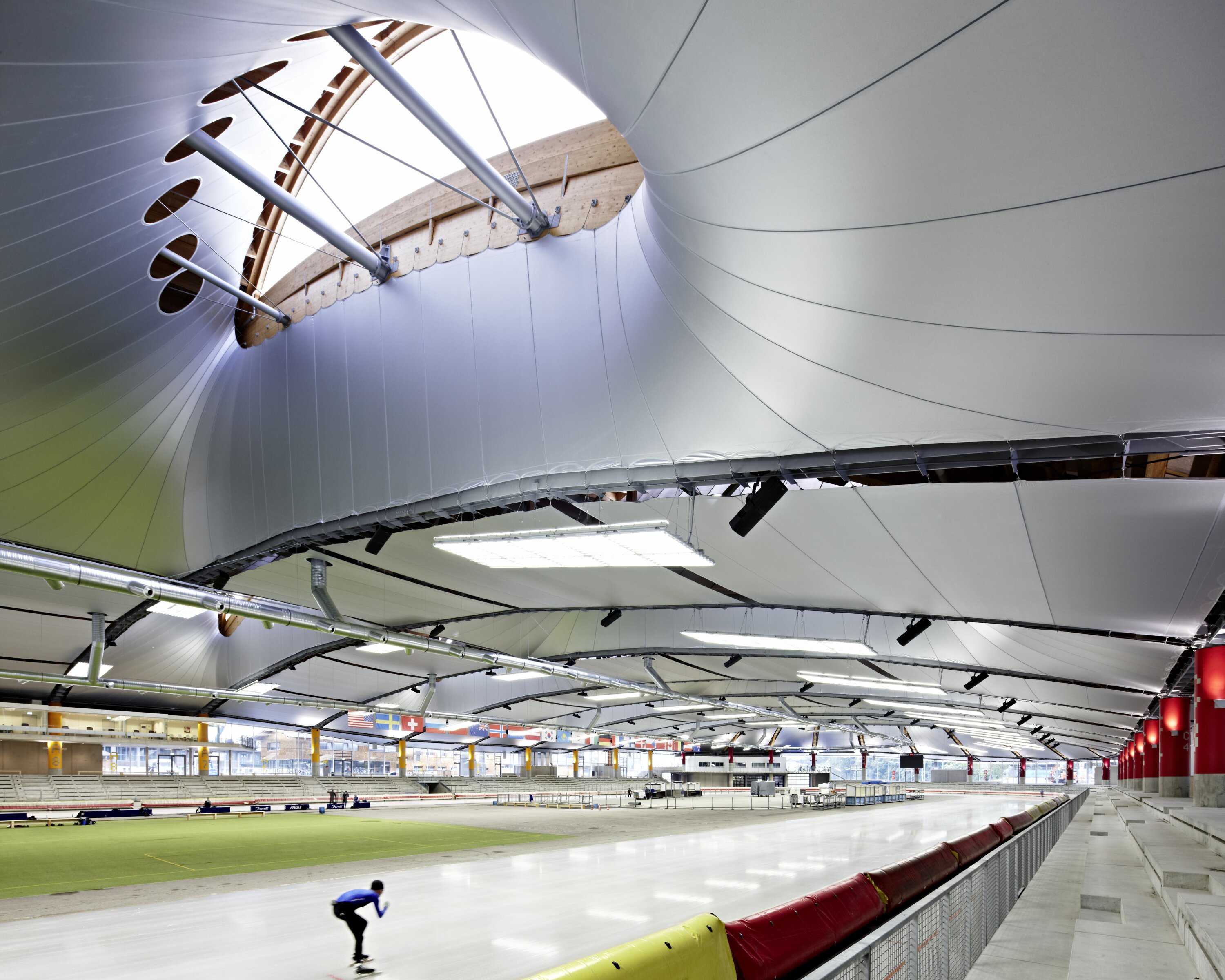 Project by Behnisch Architekturbuero, Max Aicher Arena. Wide view of an indoor ice rink with a track, a skater in motion, and a large arched roof with lights and skylights.