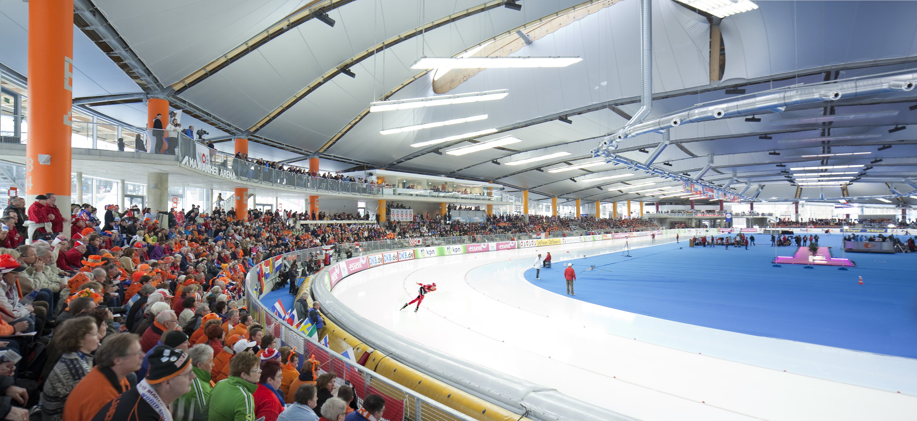Project by Behnisch Architekturbuero, Max Aicher Arena. Indoor speed skating arena with a skater racing on the track as large crowds fill tiered seating around the rink.