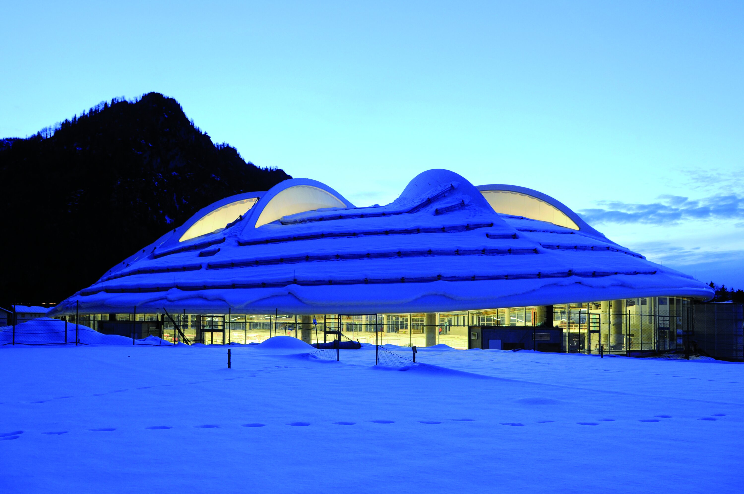 Project by Behnisch Architekturbuero, Max Aicher Arena. Snow-covered domed building with glowing windows at dusk, set in a snowy landscape with mountains in the background.