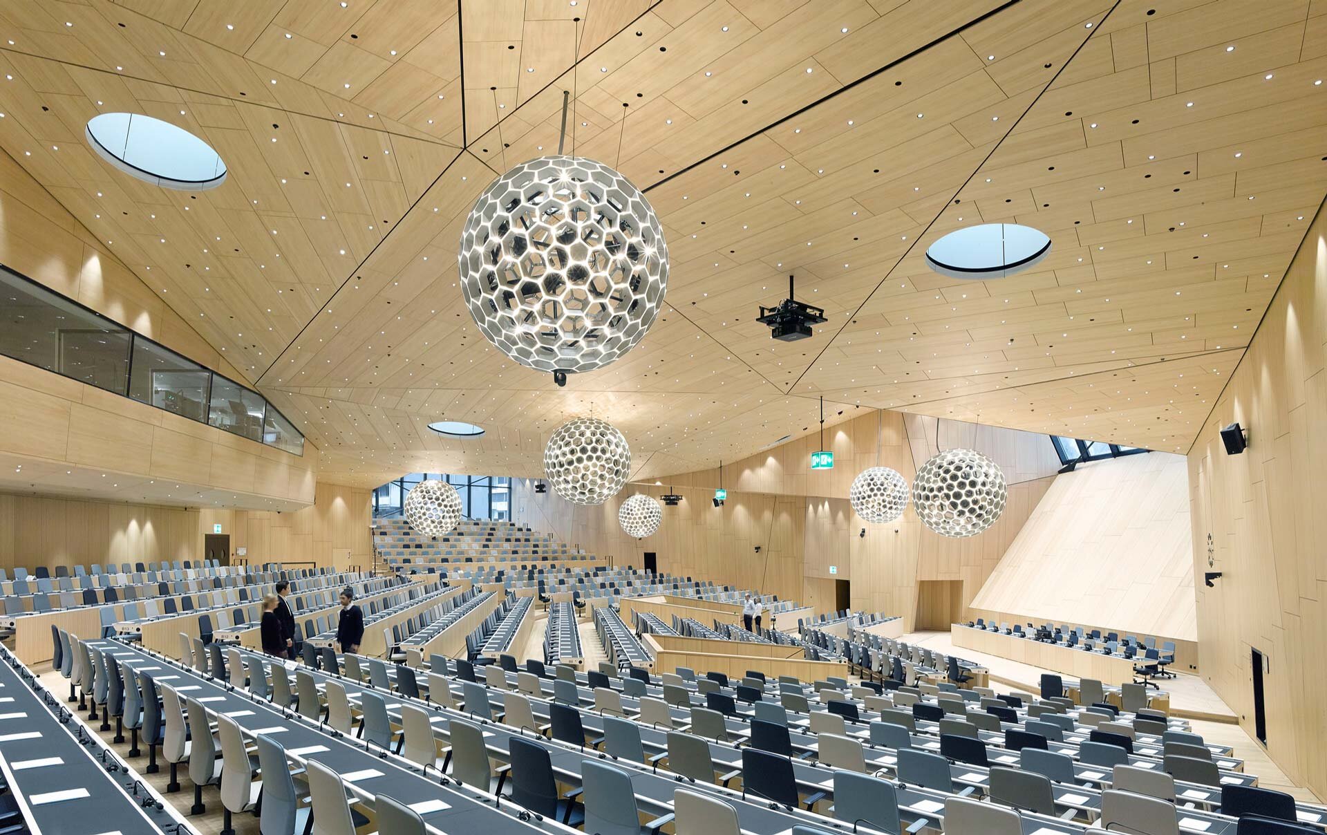 Project by Behnisch Architekturbuero, WIPO Conference Hall. Large lecture hall with tiered seating, wood-paneled walls and ceiling, and spherical pendant lights hanging above rows of desks.