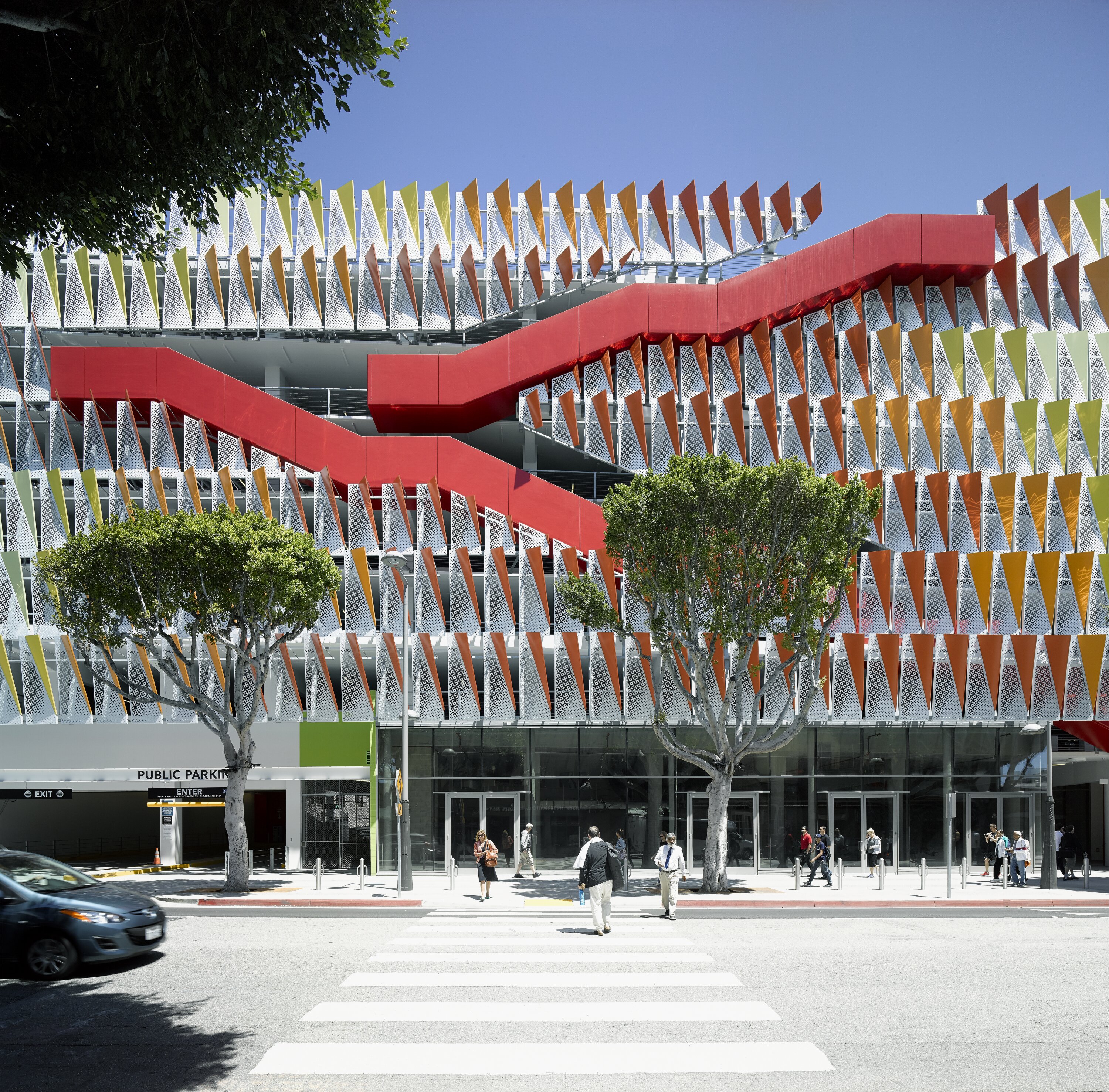 Project by Behnisch Architekturbuero, City of Santa Monica Parking Structure #6. Front view of the building. The patterned facade and the striking red exterior staircase, which zigzags across the levels, are clearly visible. In the foreground, trees and pedestrians crossing a zebra crossing can be seen.