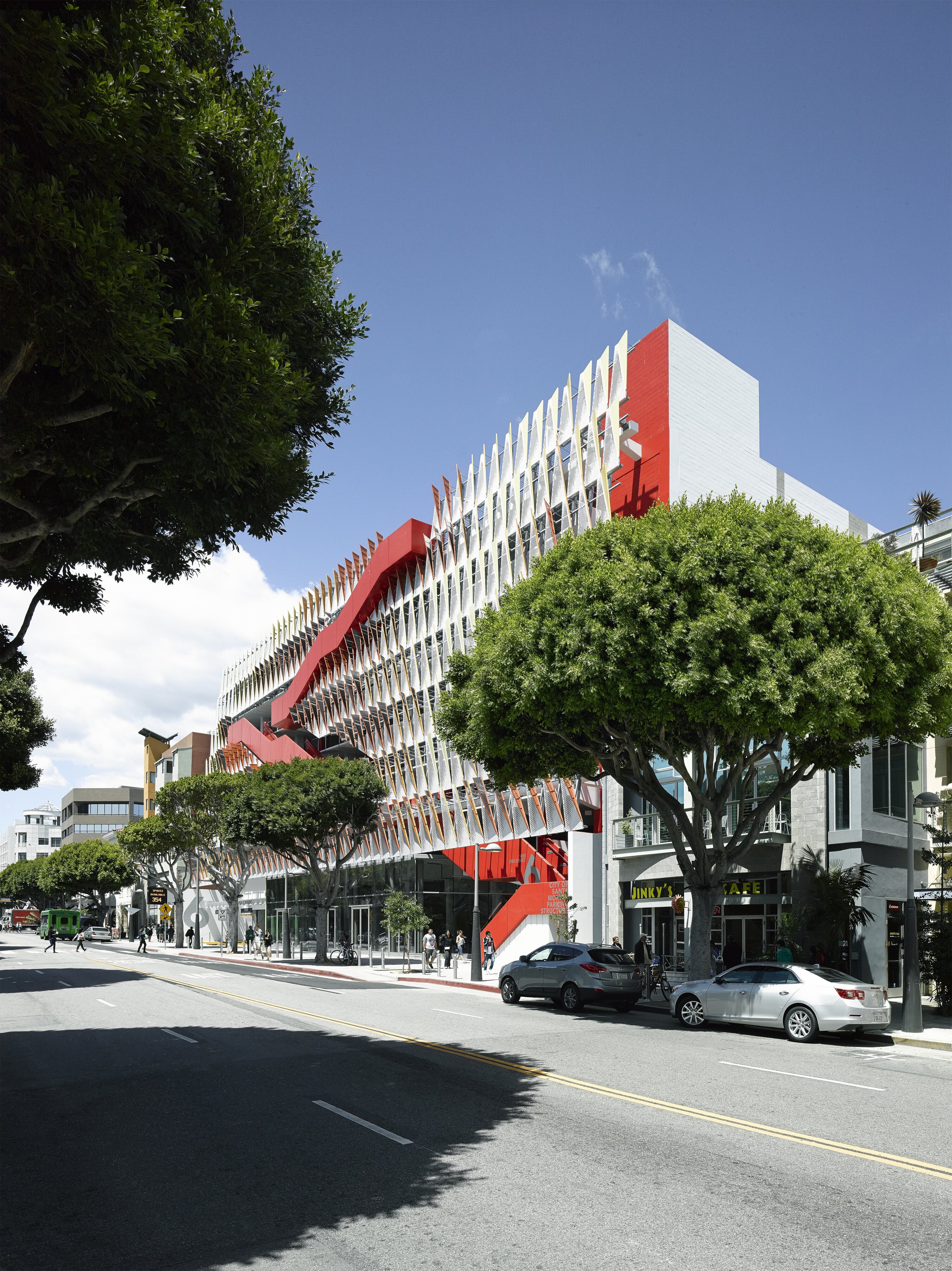 Project by Behnisch Architekturbuero, City of Santa Monica Parking Structure #6. The parking garage with patterned facade and bold red exterior stair zigzagging across levels can be seen in the background. Trees are hiding the building and are in front of it.