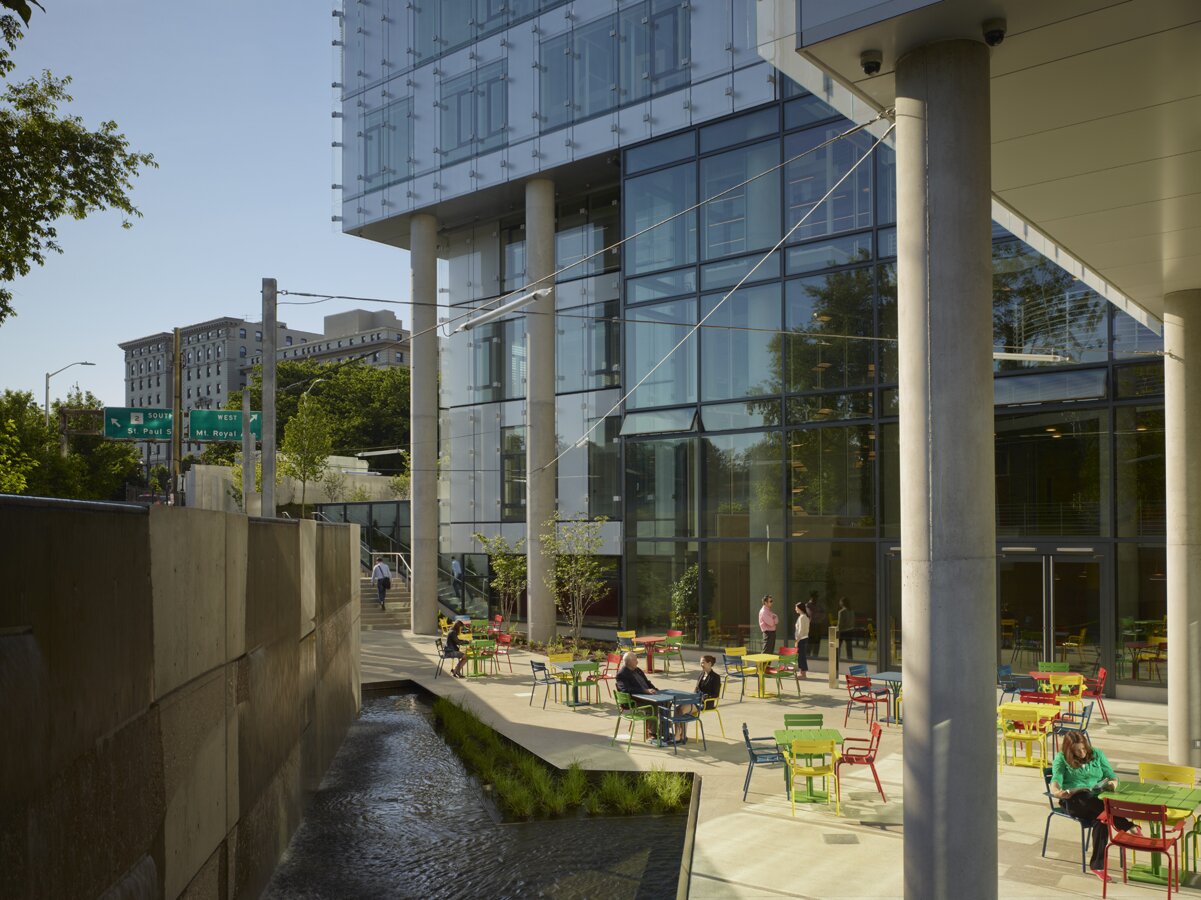 Project by Behnisch Architekturbuero, The John and Frances Angelos Law Center. An outdoor terrace, supported by columns, adjoins the atrium area. People are sitting at brightly colored tables and chairs. A stream runs alongside the terrace.