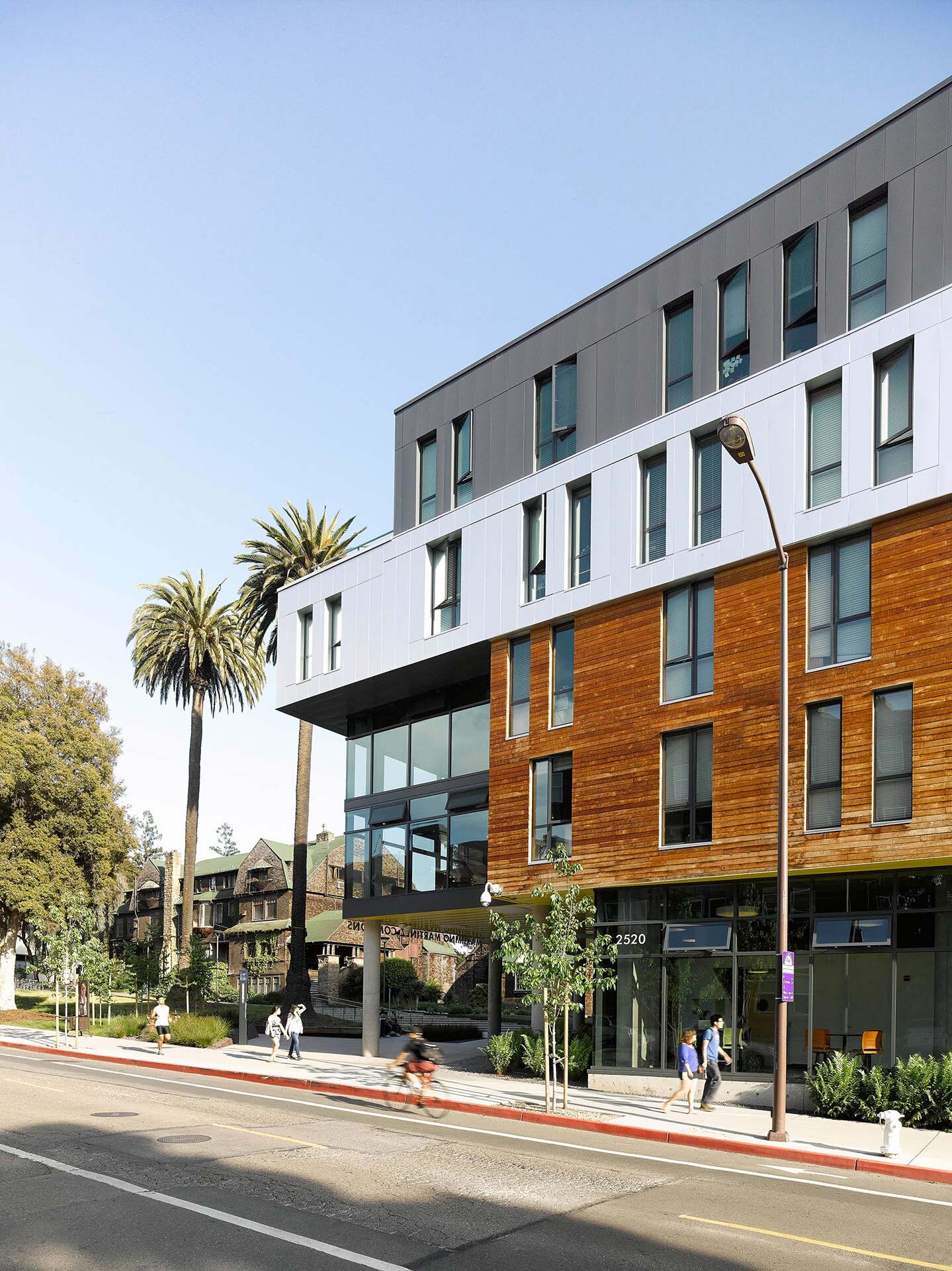 Project by Behnisch Architekturbuero, Maximino Martinez Commons, University of California. Modern multi-story building with white and wood facade next to a street. Pedestrians walk on sidewalks and a cyclist rides past in the foreground.