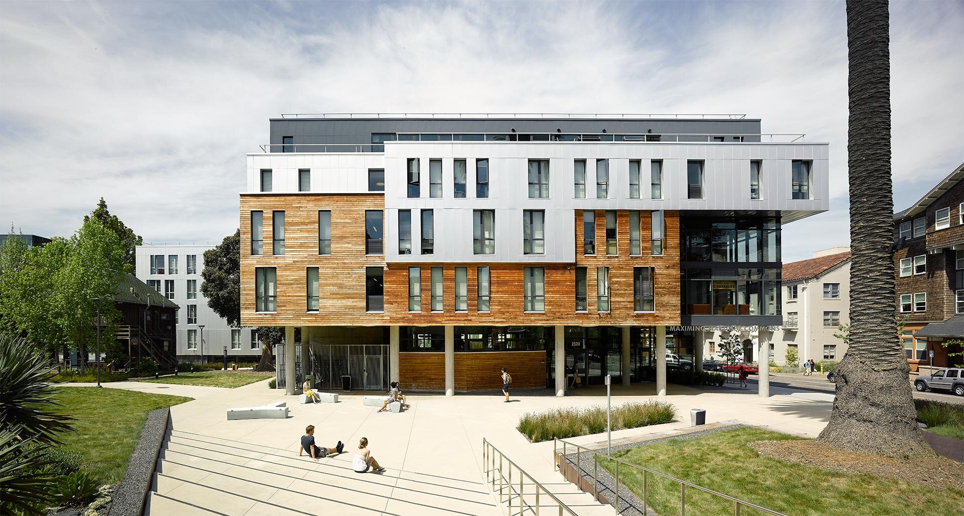 Project by Behnisch Architekturbuero, Maximino Martinez Commons, University of California. Modern multi-story building with a white and wooden facade on stilts, open square with stairs and seating, and people walking or sitting outdoors.