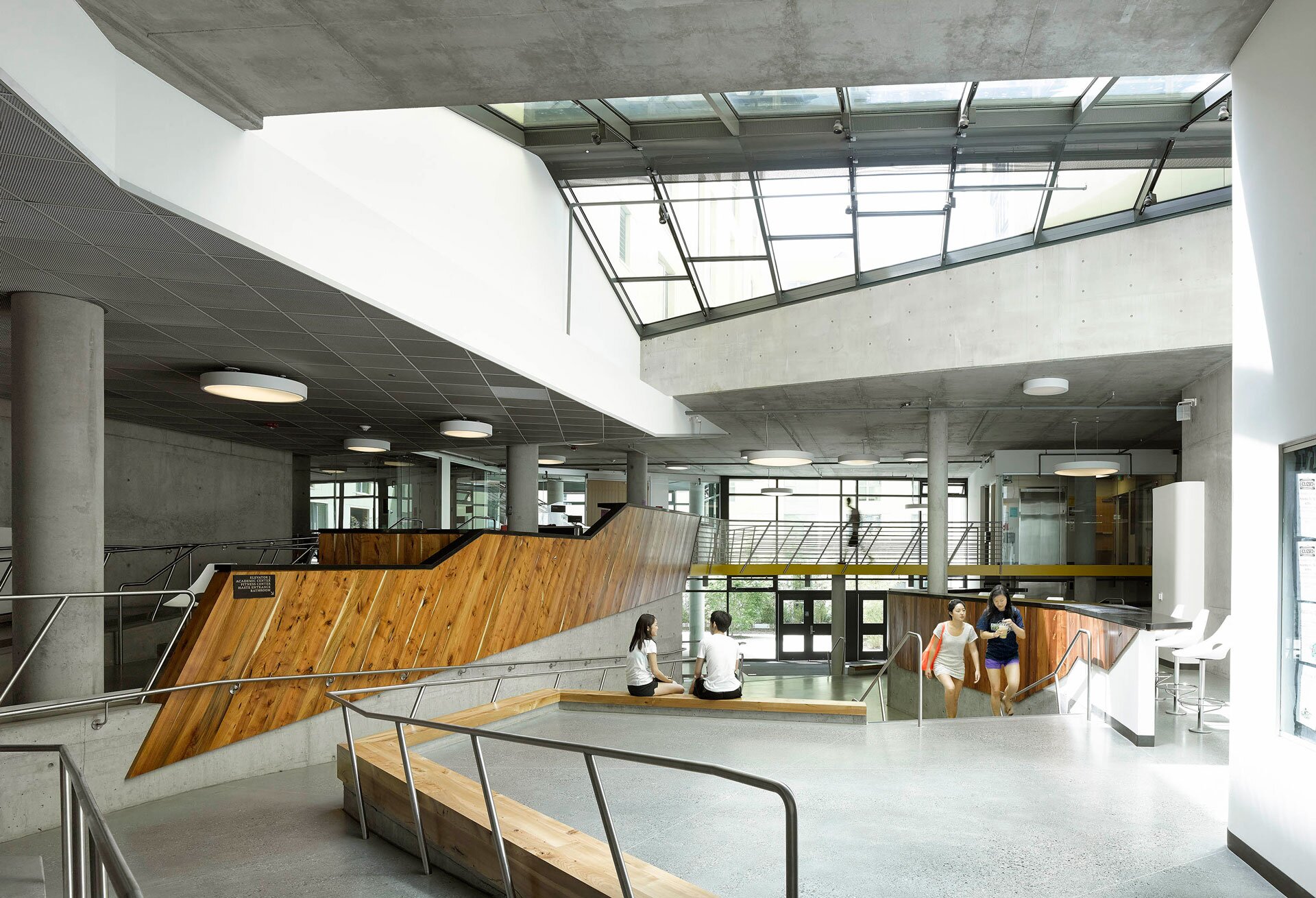 Project by Behnisch Architekturbuero, Maximino Martinez Commons, University of California. An open atrium with concrete columns, a skylight, and wood-trimmed ramps. People sit, walk, and converse on stairs and across several levels inside.