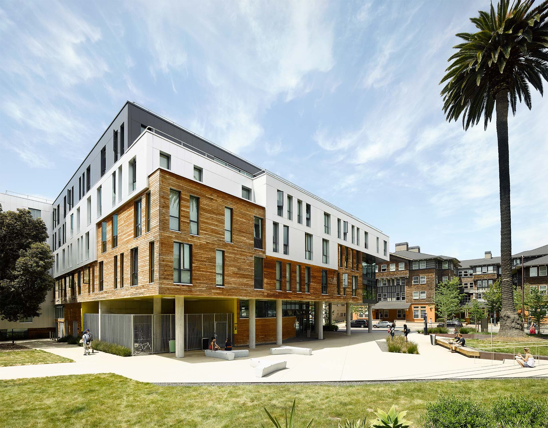 Project by Behnisch Architekturbuero, Maximino Martinez Commons, University of California. Modern multi-story building with a white and wooden facade on stilts, open square with stairs and seating, and people walking or sitting outdoors.