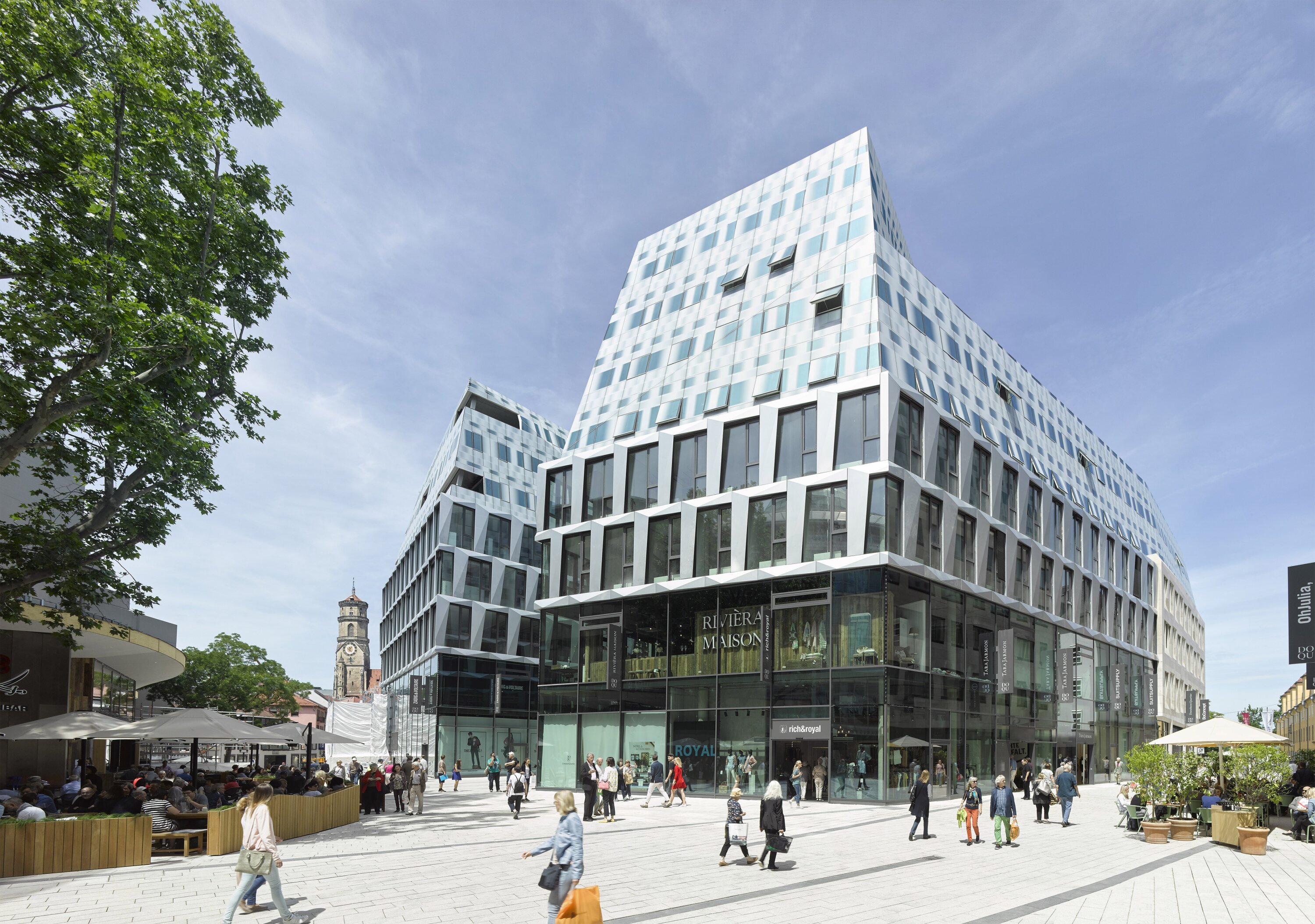 Project by Behnisch Architekturbuero, Dorotheen Quartier. Modern glass and white office building on a busy plaza with pedestrians, shops at ground level, and outdoor caf&eacute; seating nearby.