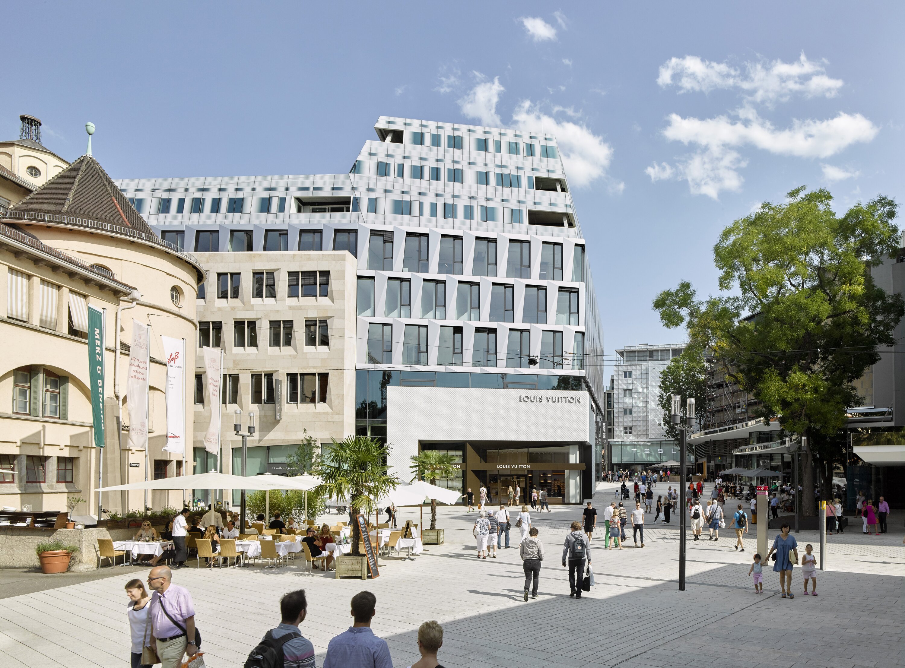 Project by Behnisch Architekturbuero, Dorotheen Quartier. Busy city square with pedestrians, outdoor caf&eacute; seating, and modern multi-story building with retail storefronts under a blue sky.