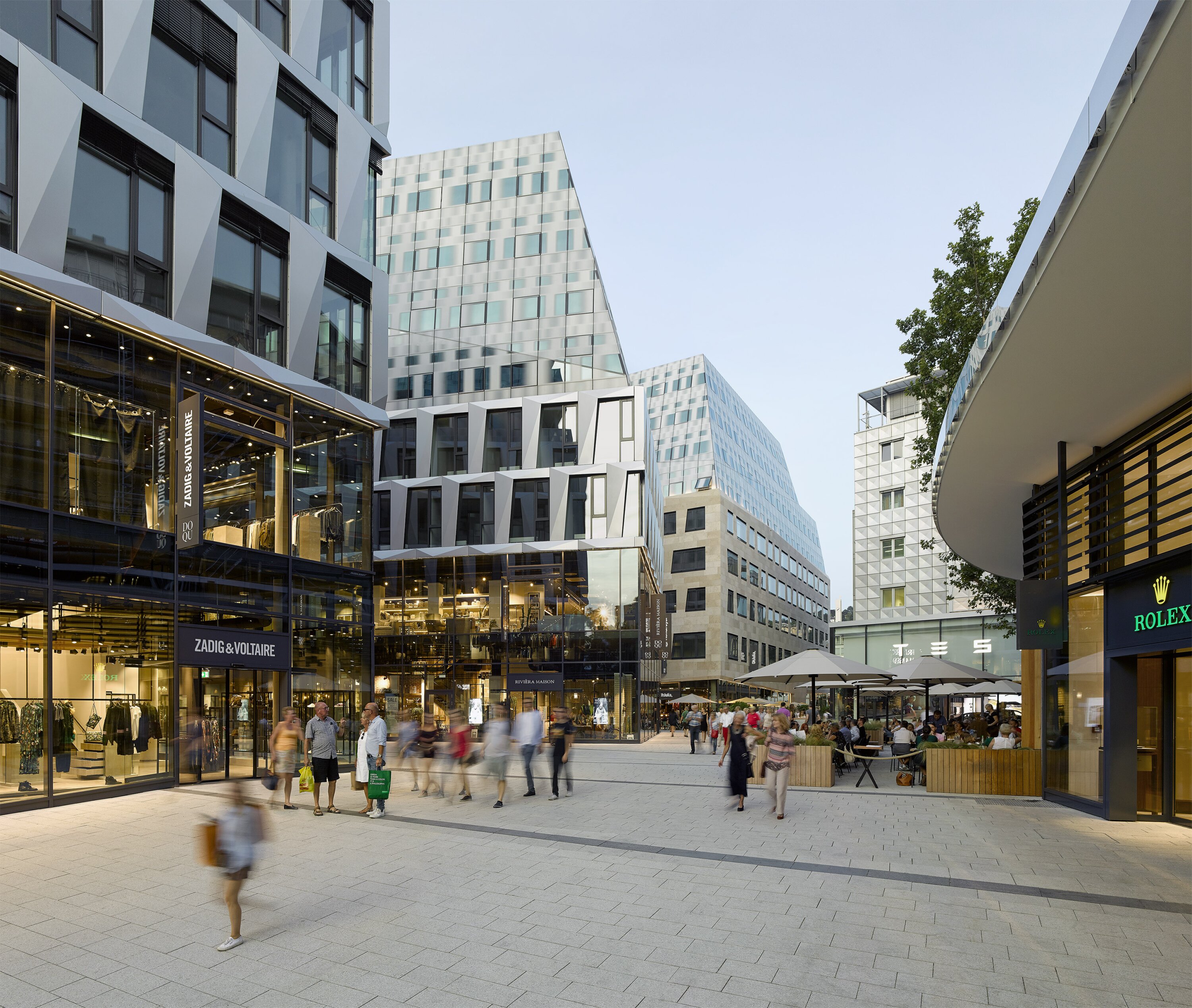 Project by Behnisch Architekturbuero, Dorotheen Quartier. City square with pedestrians, outdoor caf&eacute; seating, and the multi-story building with retail storefront in an evening atmosphere.