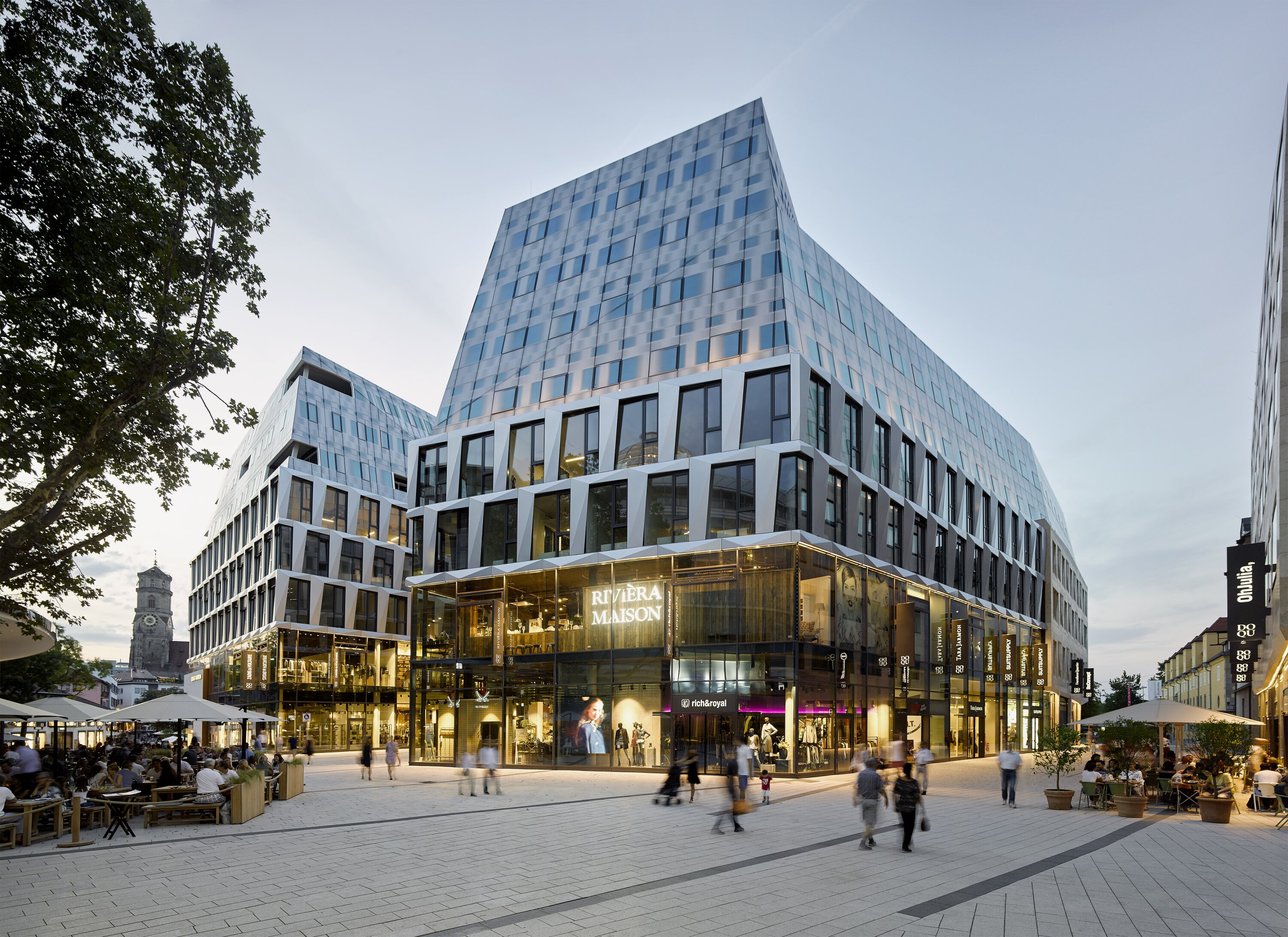 Project by Behnisch Architekturbuero, Dorotheen Quartier. City square with pedestrians, outdoor caf&eacute; seating, and the multi-story building with retail storefront in an evening atmosphere.