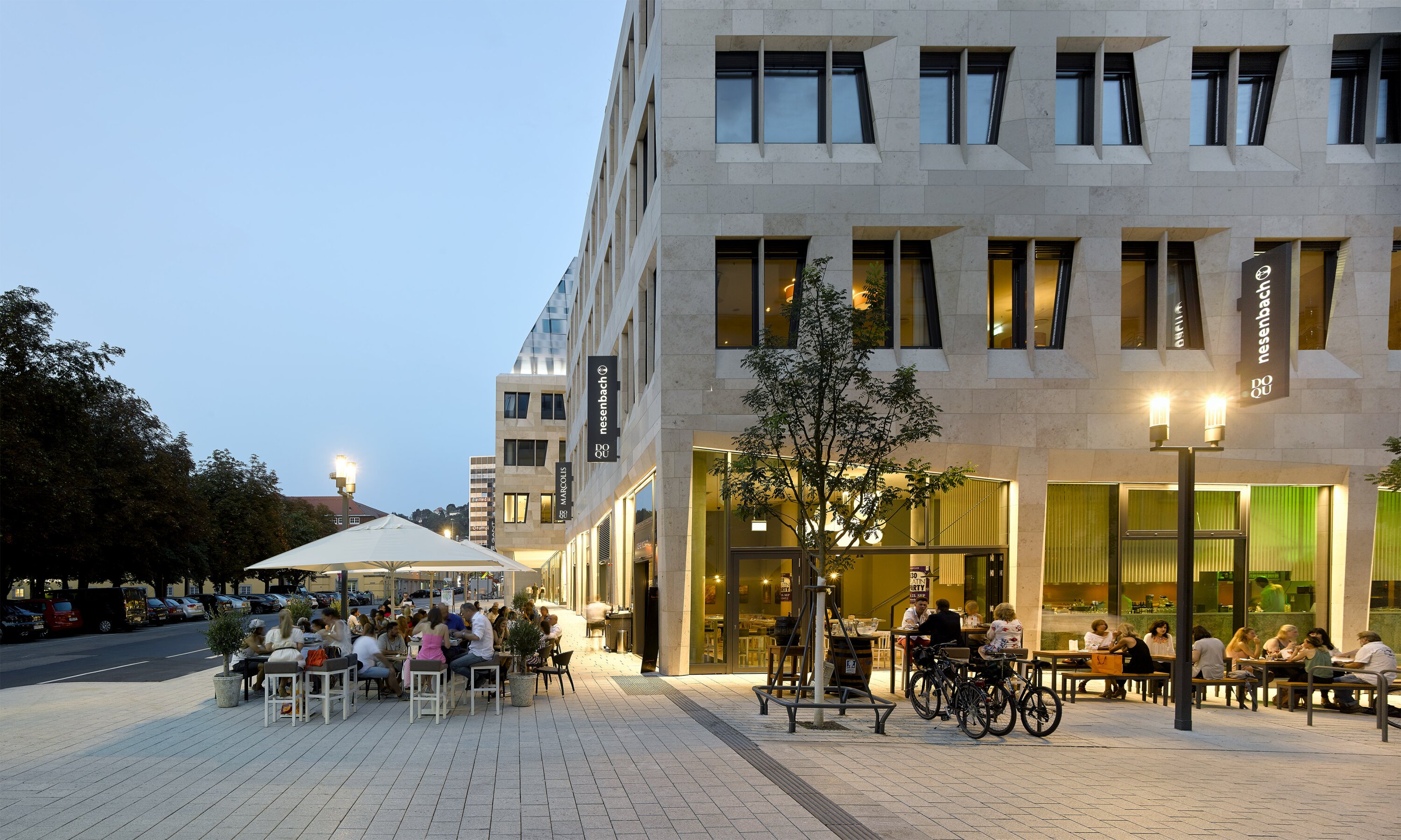 Project by Behnisch Architekturbuero, Dorotheen Quartier. Evening street scene with outdoor caf&eacute; seating beside the building, people dining, bicycles parked, and warm interior lights glowing.